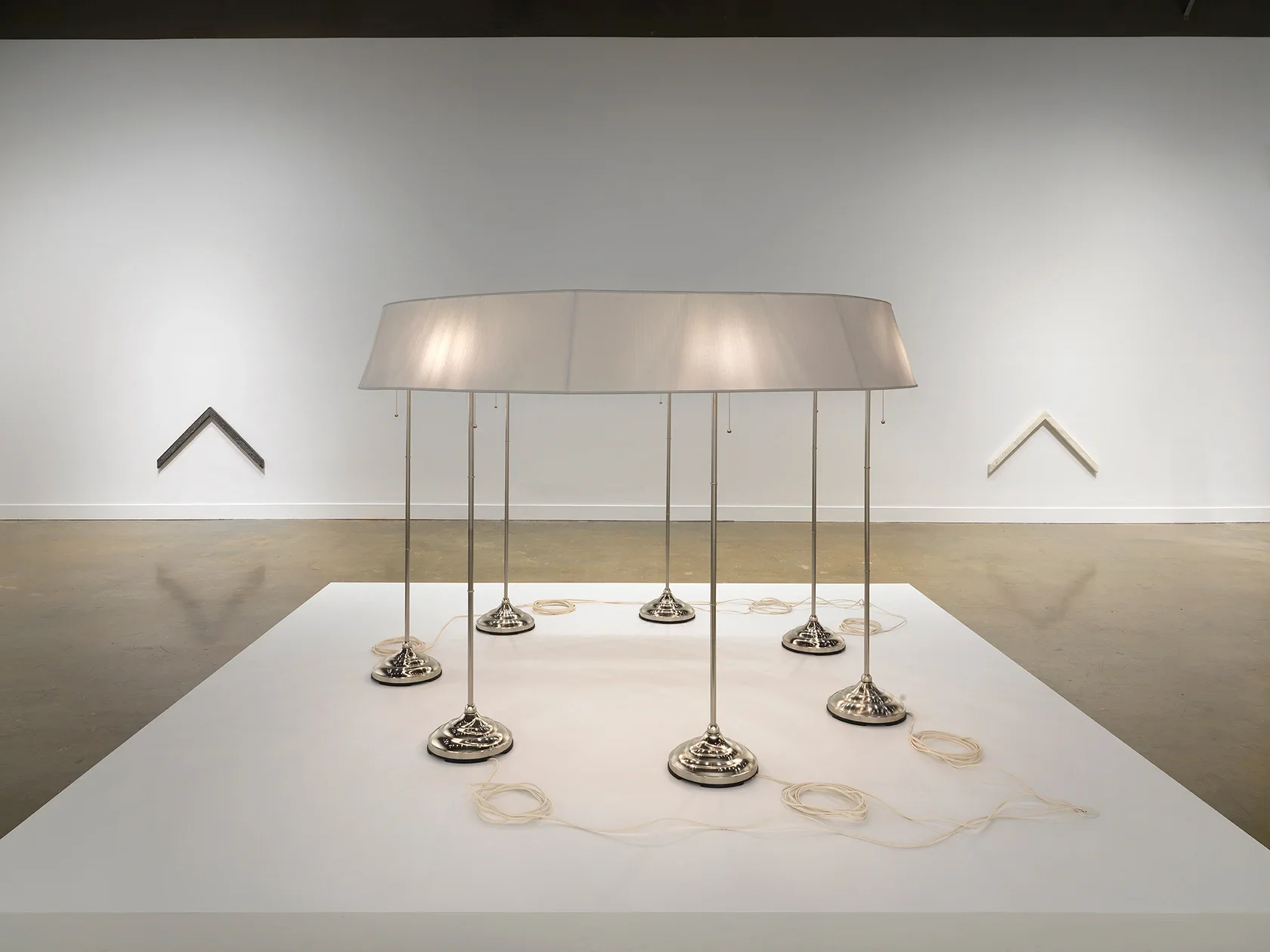 On a low pedestal in the foreground, seven standing lamps are conjoined by a single ring-shaped lampshade. A neat coil of pale cord sits near the base of each lamp. The sculpture is flanked, in the background, by two chevron-shaped wall pieces. The one on the left is dark, while the one on the right is pale.