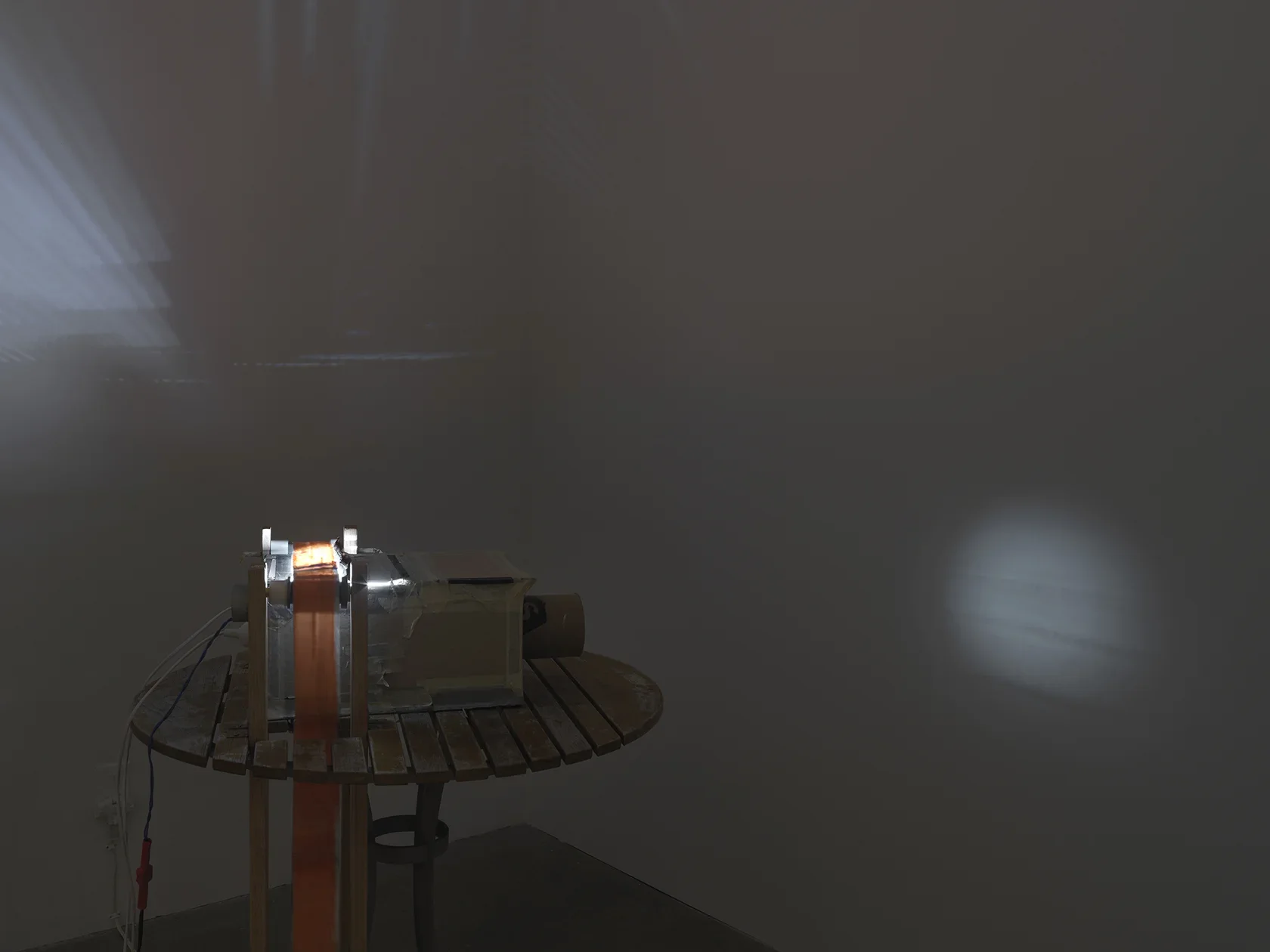 A boxy makeshift projector sits on a wooden table; it is crudely constructed with materials like cardboard and duct tape. Three cords are attached to its back, and a strip of semi-transparent material runs through the device, like a film strip would. A circle of light with a blurry strip of text is projected through the projector’s cylindrical “lens.” A more abstract pattern of light rays is also thrown across an adjacent wall.