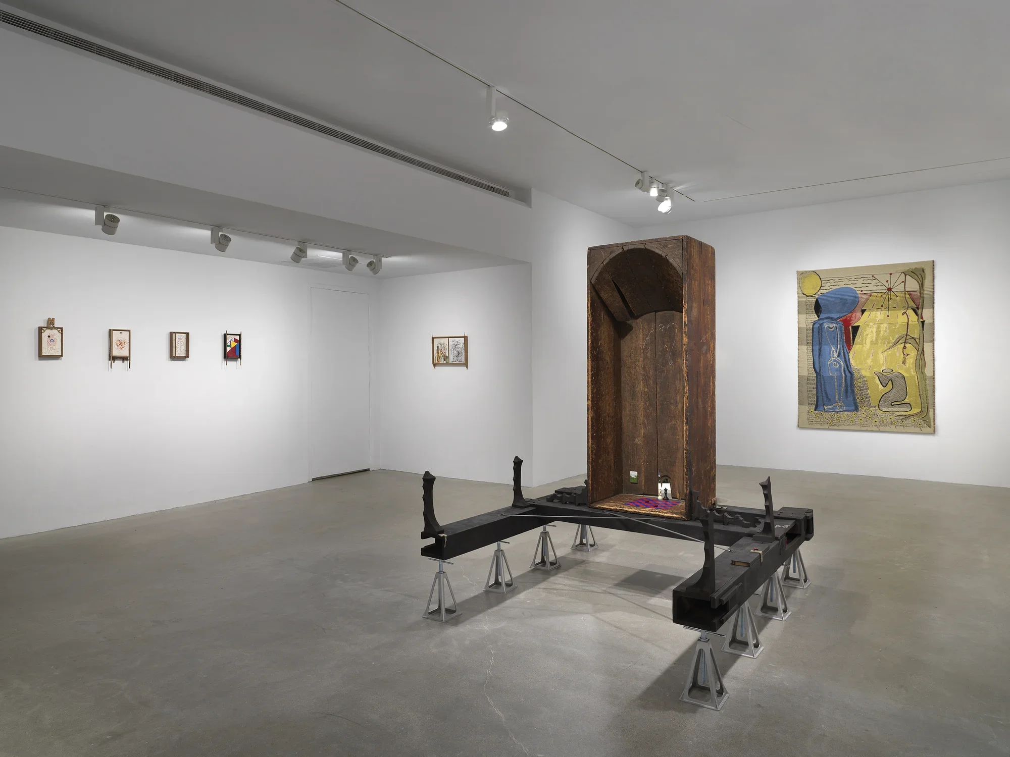 In the foreground, a vertical wooden bathtub sits on horizontal wooden beams, which are held up by eight metal supports. Inside the bathtub are small painted scenes and a red and blue checkerboard pattern on the base. On the left walls, five small drawings hang in wooden frames. In the rear, a golden tapestry depicts two figures, one standing and one kneeling.