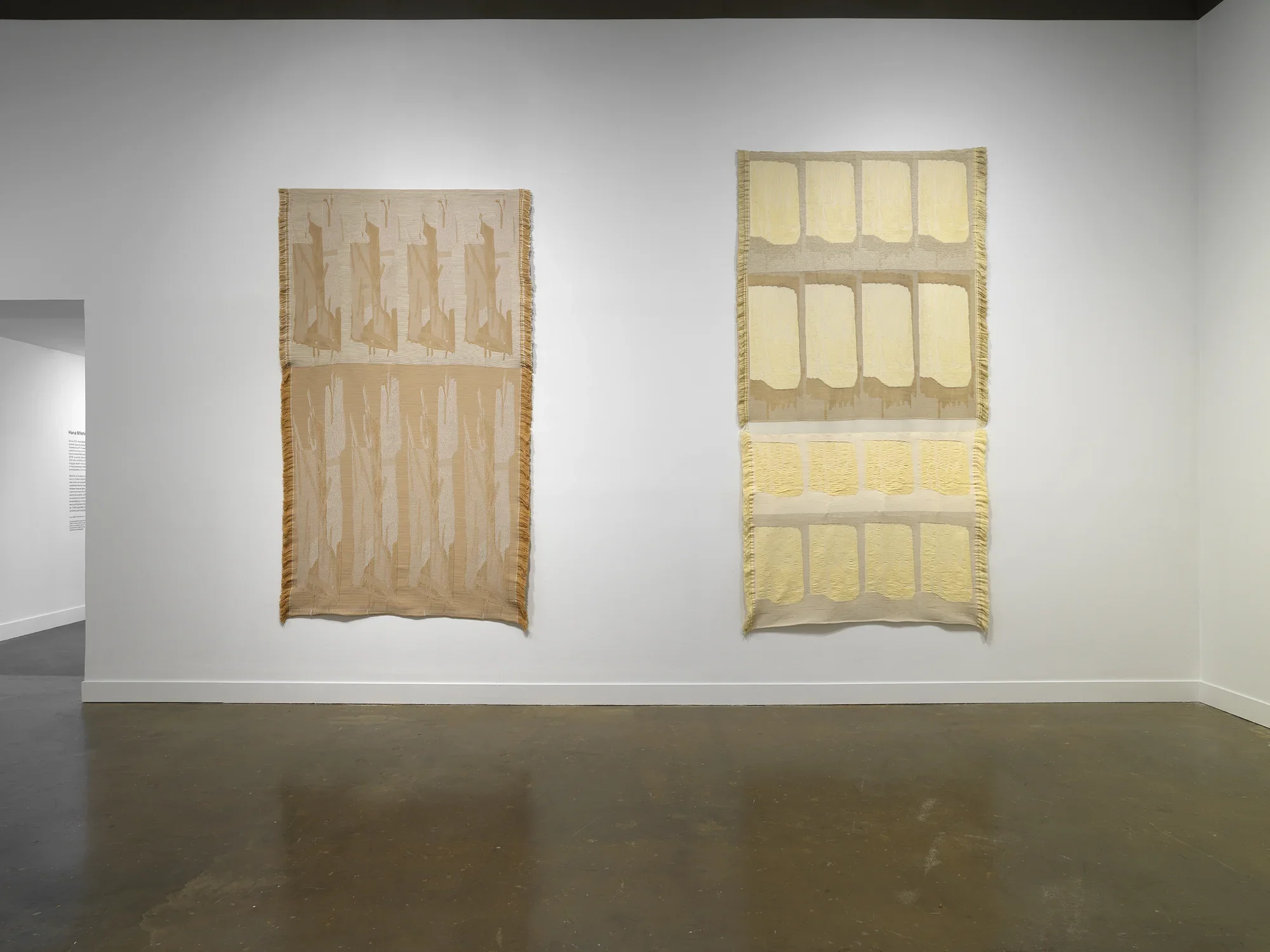 Two tapestries, similar in scale and color, hang side by side. Each depicts abstracted forms that are repeated in a grid with slight variations, as if some rows were stretched. They are brown, beige, yellow, and gold.