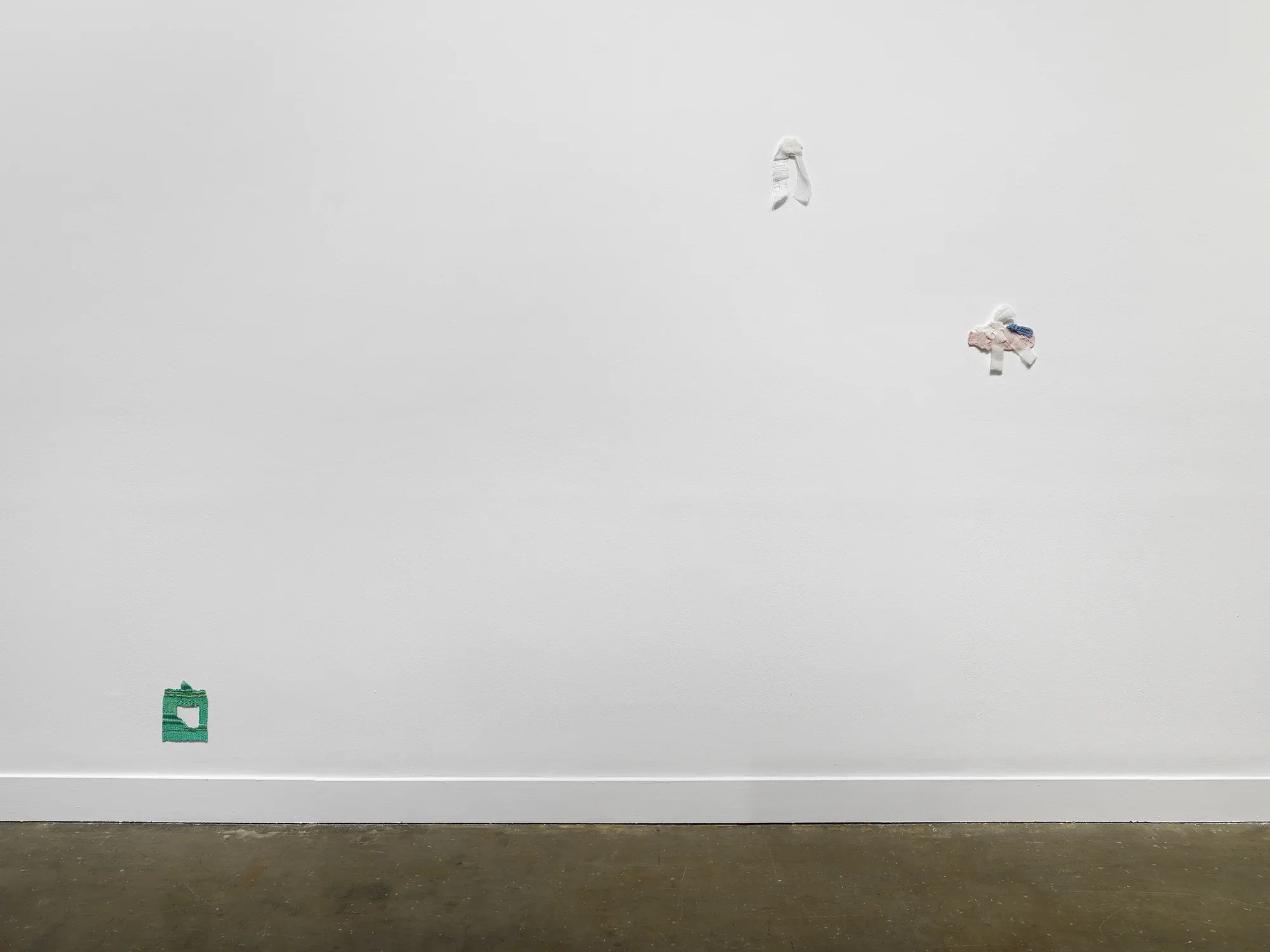 Three small fiber artworks hang at various heights on a white wall. A small green woven square with the center peeled back hangs low to the ground on the left. Two works hang in the upper right corner: one is a white and ribbon-like; the other is an overlapping bundle of pink, white, and blue textile.