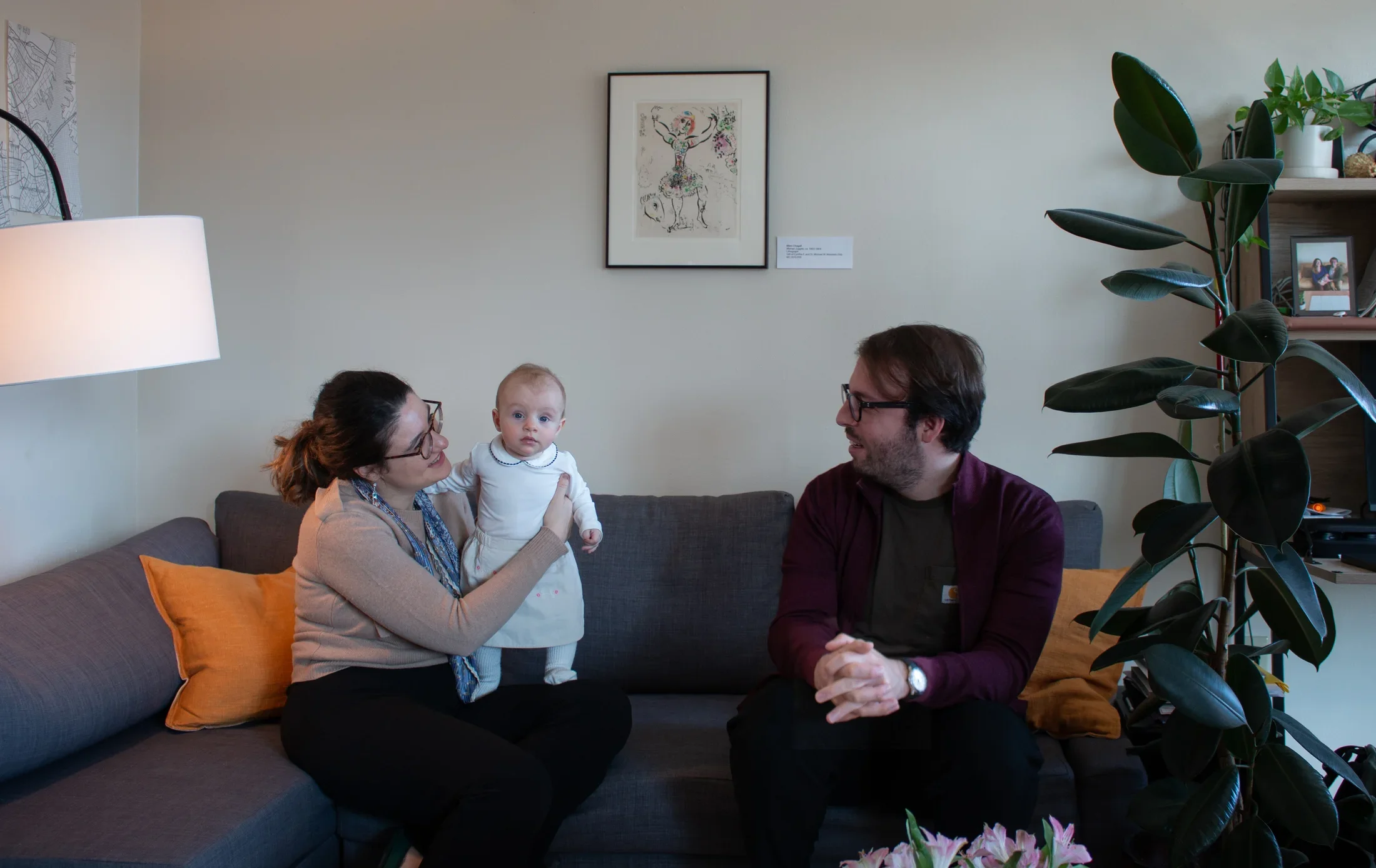 A man and woman holding a baby are sitting on a gray couch with a framed artwork on the wall behind.