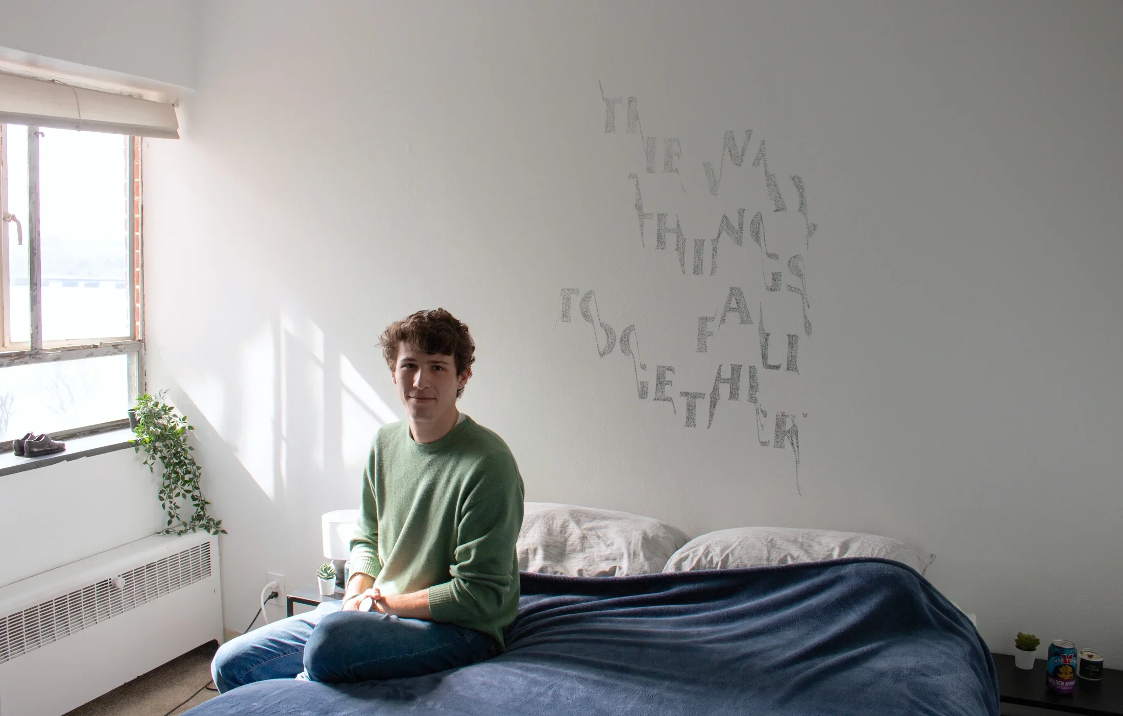 A man wearing a green sweater is sitting on a bed with navy sheets in front of a stenciled artwork on the white wall behind. The bed is to the right of a window with a plant on the windowsill.