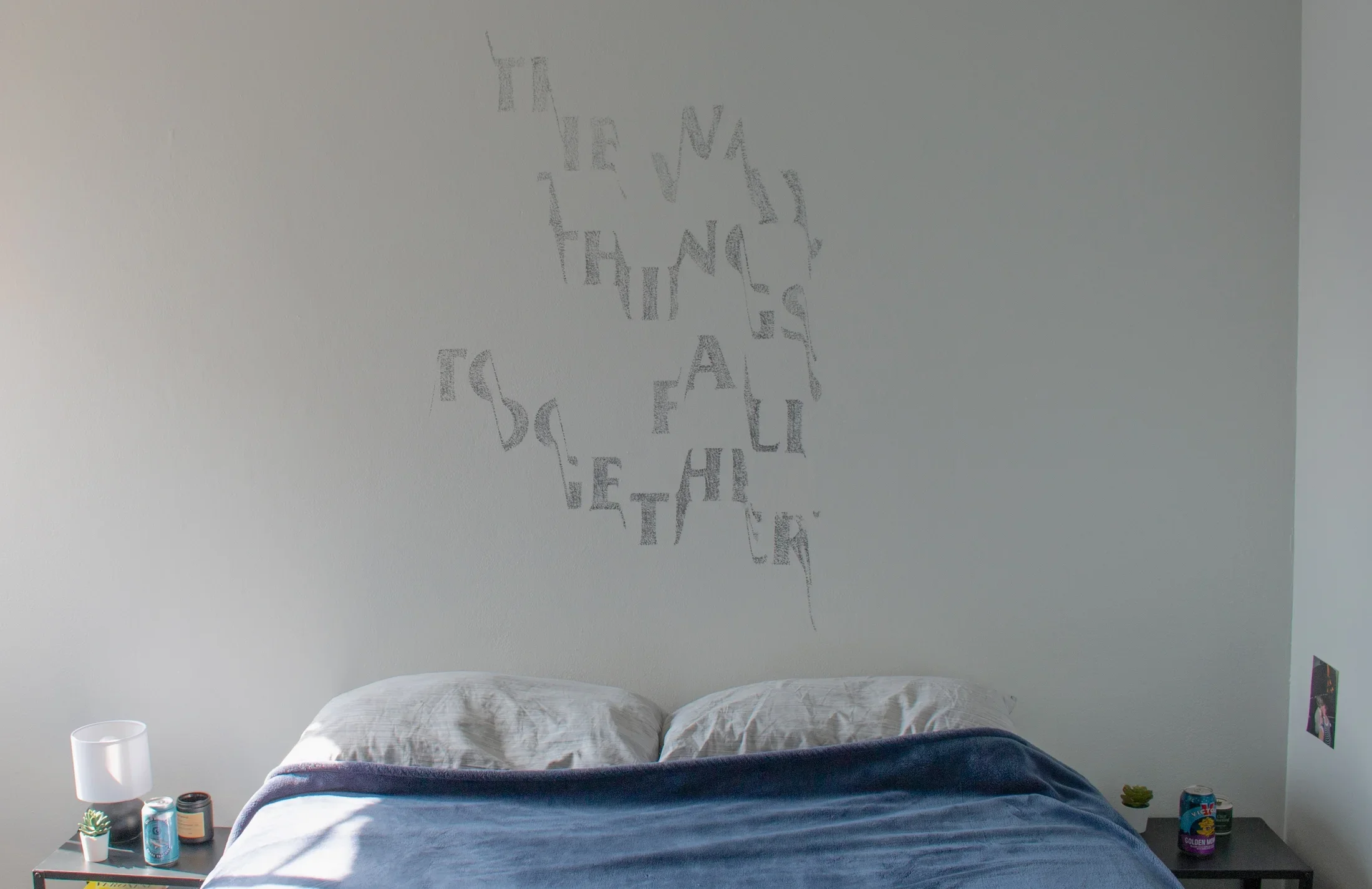 A bed with navy sheets in front of a stenciled artwork on the white wall behind. One beer can is on top of a black nightside table on either side of the bed.