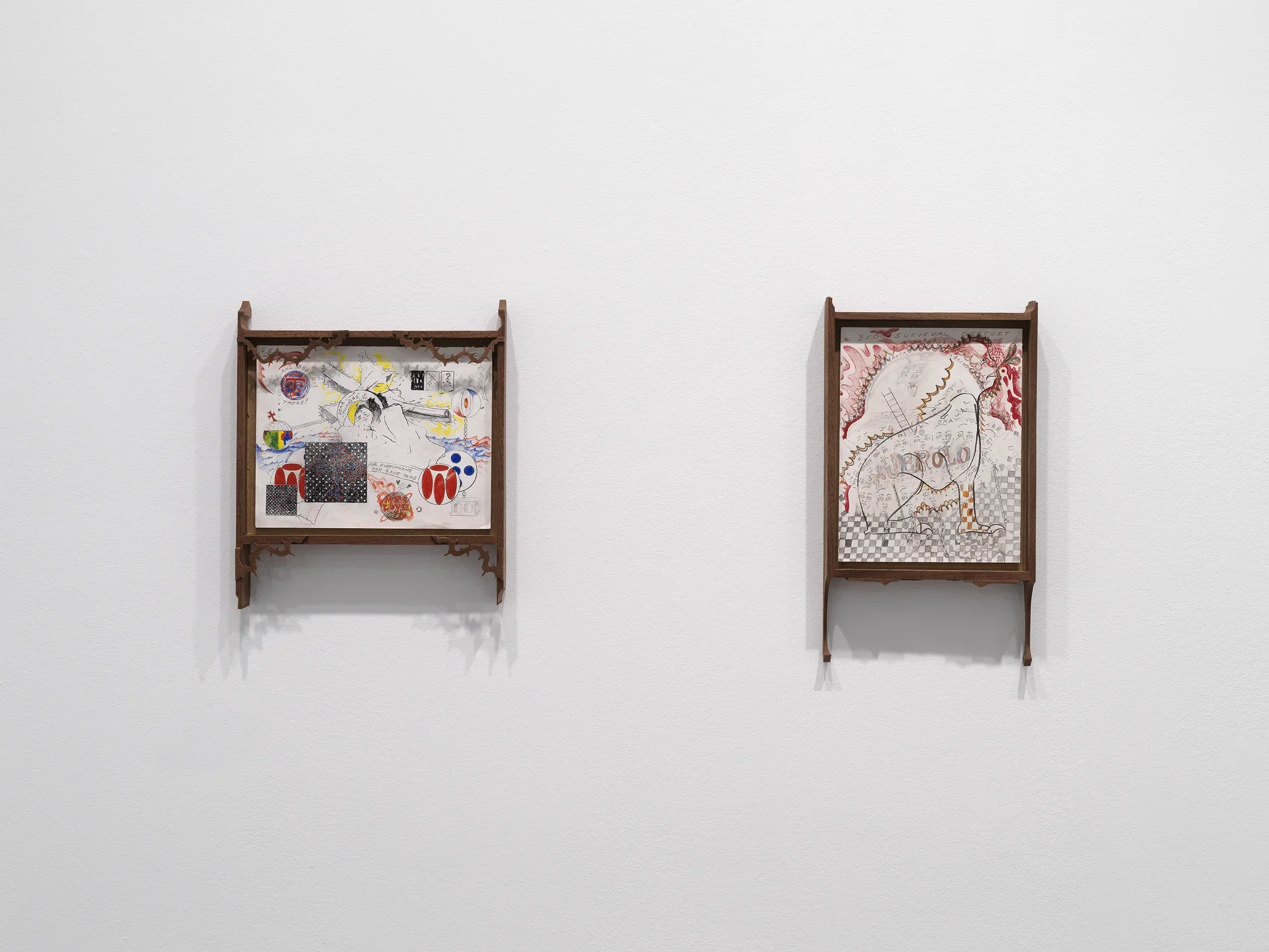 On a white wall hang two drawings in wooden frames. The left drawing is horizontal and has colorful abstract forms scattered across the paper, including forms that resemble globes, playing cards, and waves. The wooden frame is adorned with thorn motifs in each corner. The right vertical drawing depicts a four-legged animal-like form with red flame-like designs in the background and a gray and white checkerboard print at the bottom.