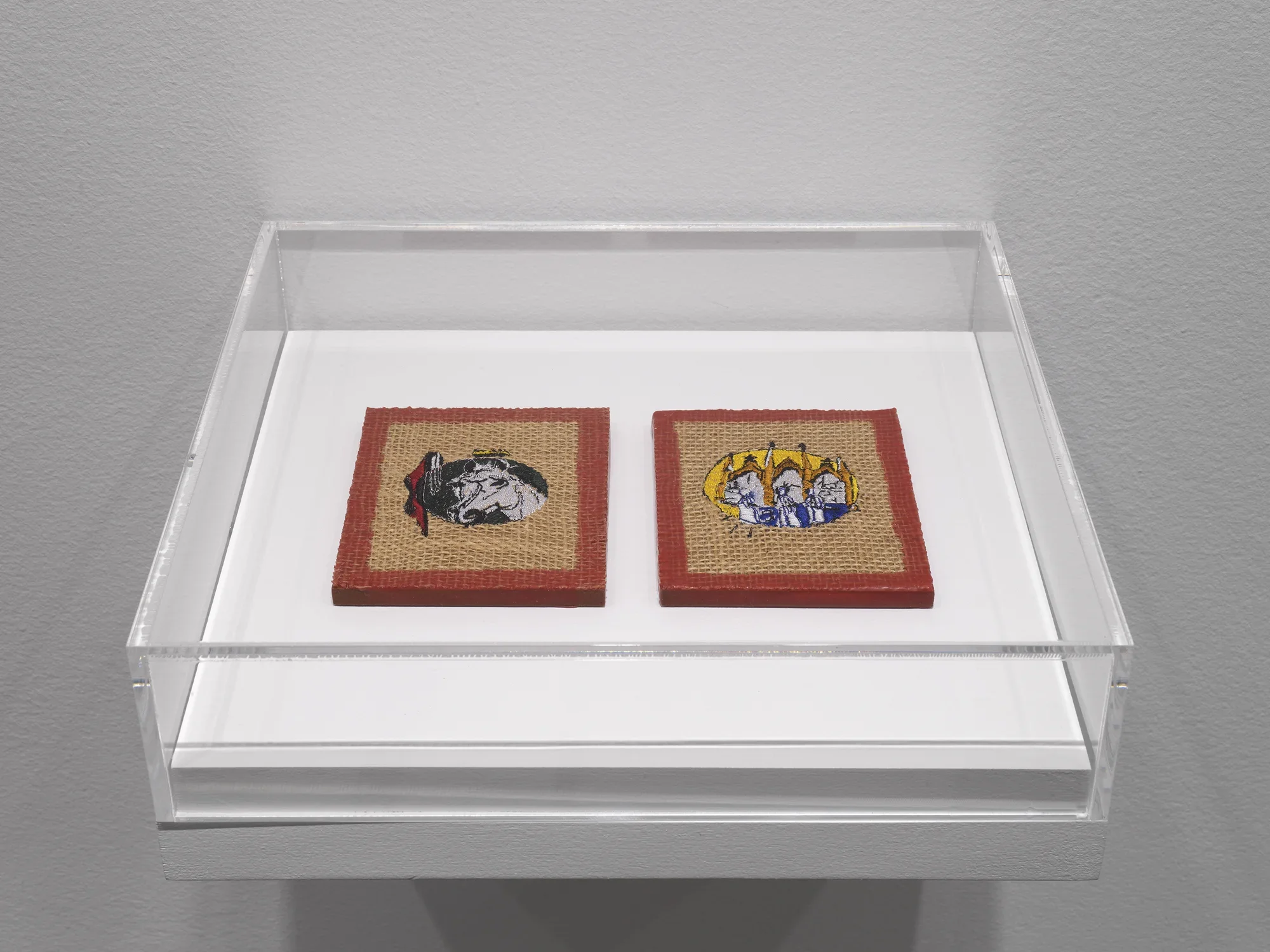 A plexiglass case hangs off of a white wall. Inside the case, two burlap-like square artworks are adorned with embroidered details. The left shows the decorated head of a horse, while the left is more ambiguous but has a similar medieval flavor. Each square has a border of red wax around the piece.