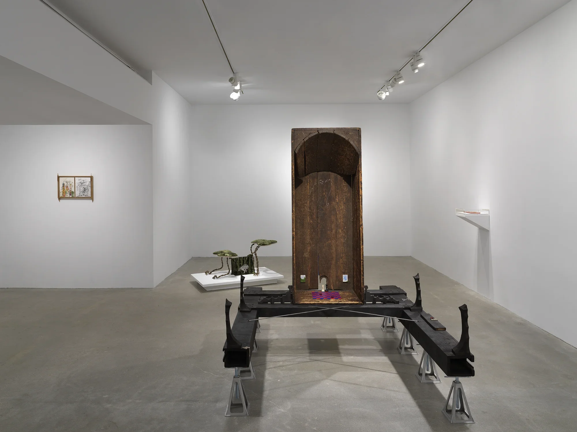 In the center of the room, a vertical wooden bathtub sits on horizontal wooden beams, which are held up by eight metal supports. Inside the bathtub are small painted scenes and a red and blue checkerboard pattern on the base. On the left wall, a small diptych drawing in a wooden frame hangs. In the background behind the wooden bathtub, a small creature-like sculpture made of metal and fabric sits on a low pedestal. On the right, a white shelf hangs off the wall with a plexiglass case on top.