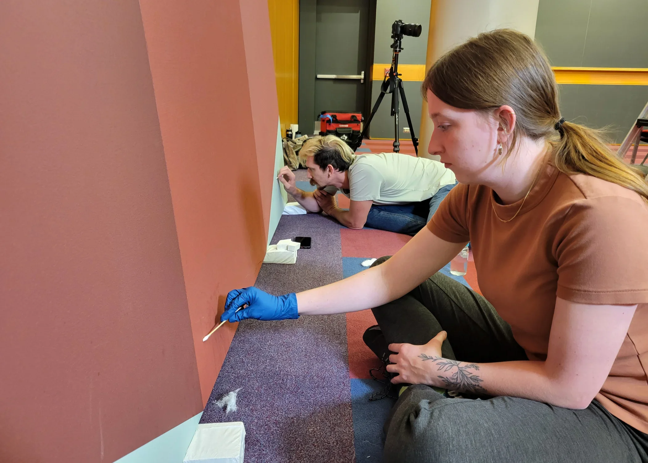 Image of two conservators sitting on the floor wearing blue gloves working on repairing a large scale red painting. 