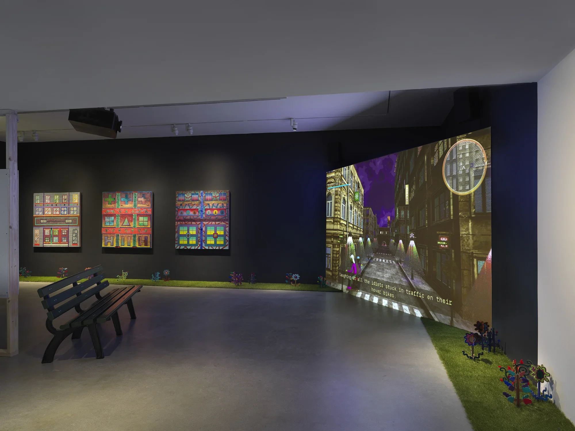 A dark gallery space with black walls is illuminated by a large screen. Digitally‬ ‭ rendered footage of a dark city street is projected onto the large screen in the right‬ ‭ corner of the room. A park bench is placed on the left side of the image facing the‬ ‭ screen. In the background, three square paintings depicting bright, geometric forms‬ ‭ hang in a row. Astroturf lines the walls of the room with painted wooden plants emerging‬ ‭ from it.‬ ‭