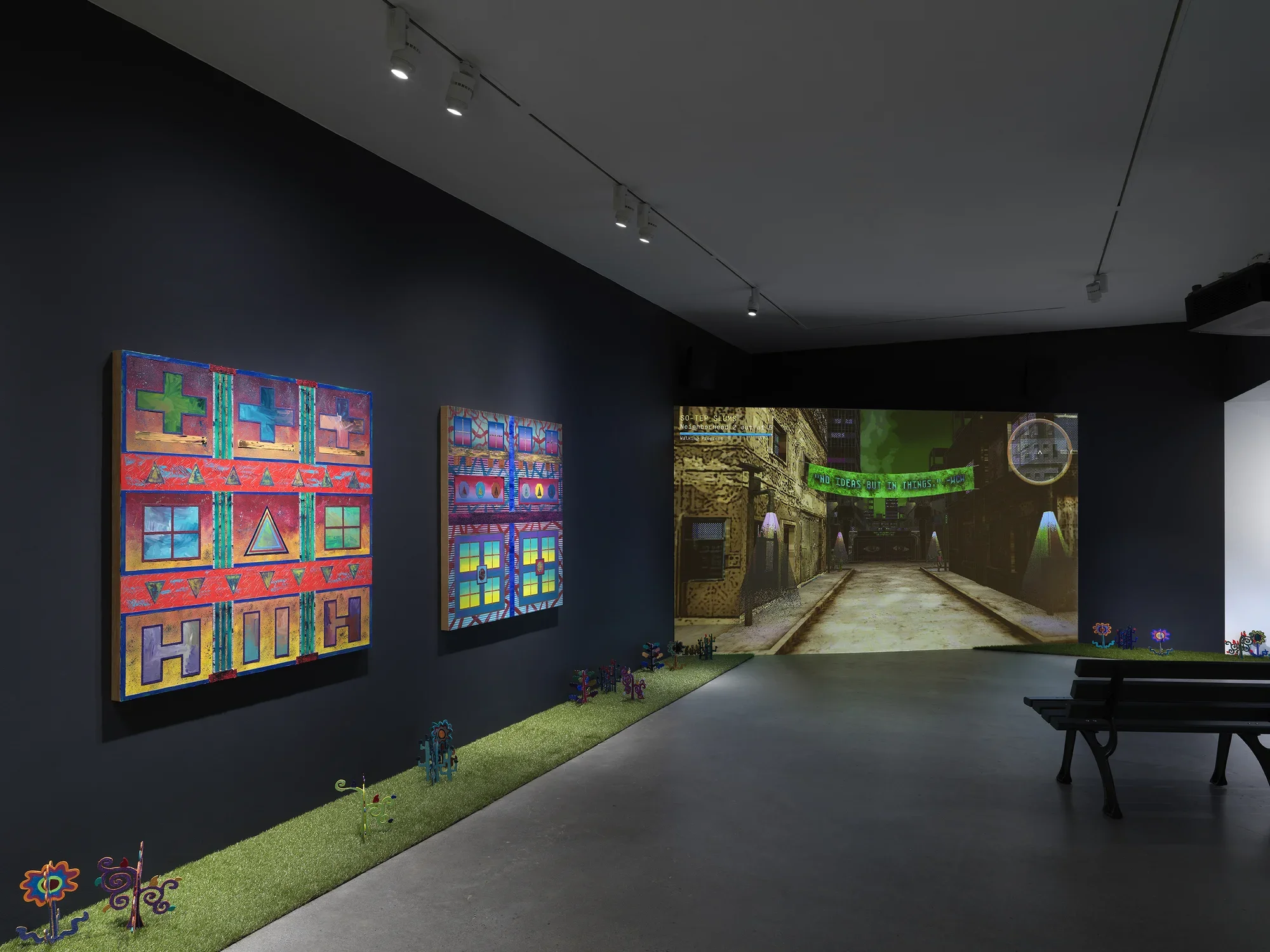 A dark, black-walled gallery space with artworks on display. On the left wall, two‬ ‭ paintings hang in a row depicting bright, geometric forms. In the back of the gallery, a‬ ‭ large screen illuminates the space displaying digitally rendered footage of a dark city‬ ‭ street with a green banner hung between buildings. A park bench faces the screen. A‬ ‭ strip of astroturf lines the floor around the gallery, with painted wooden flowers emerging‬ ‭from it.‬ ‭