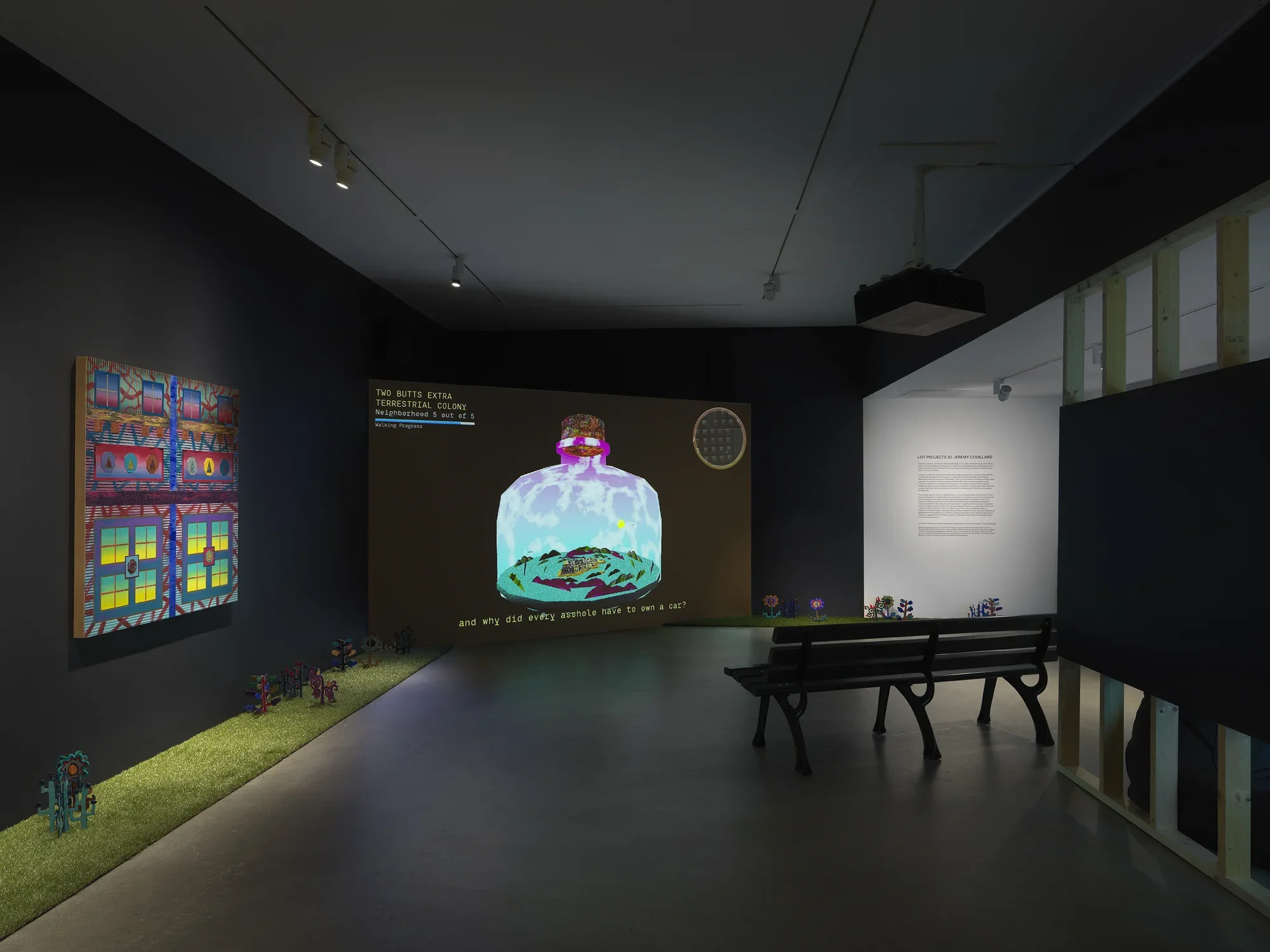 A dark, black-walled gallery space with artworks on display. On the left wall‬ ‭ hangs a painting depicting bright, geometric forms. In the back of the gallery, a large‬ ‭ screen illuminates the space displaying digitally rendered footage of a large bottle with‬ ‭ an island inside of it. A park bench faces the screen. A strip of astroturf lines the floor‬ ‭ around the gallery, with painted wooden flowers emerging from it.‬