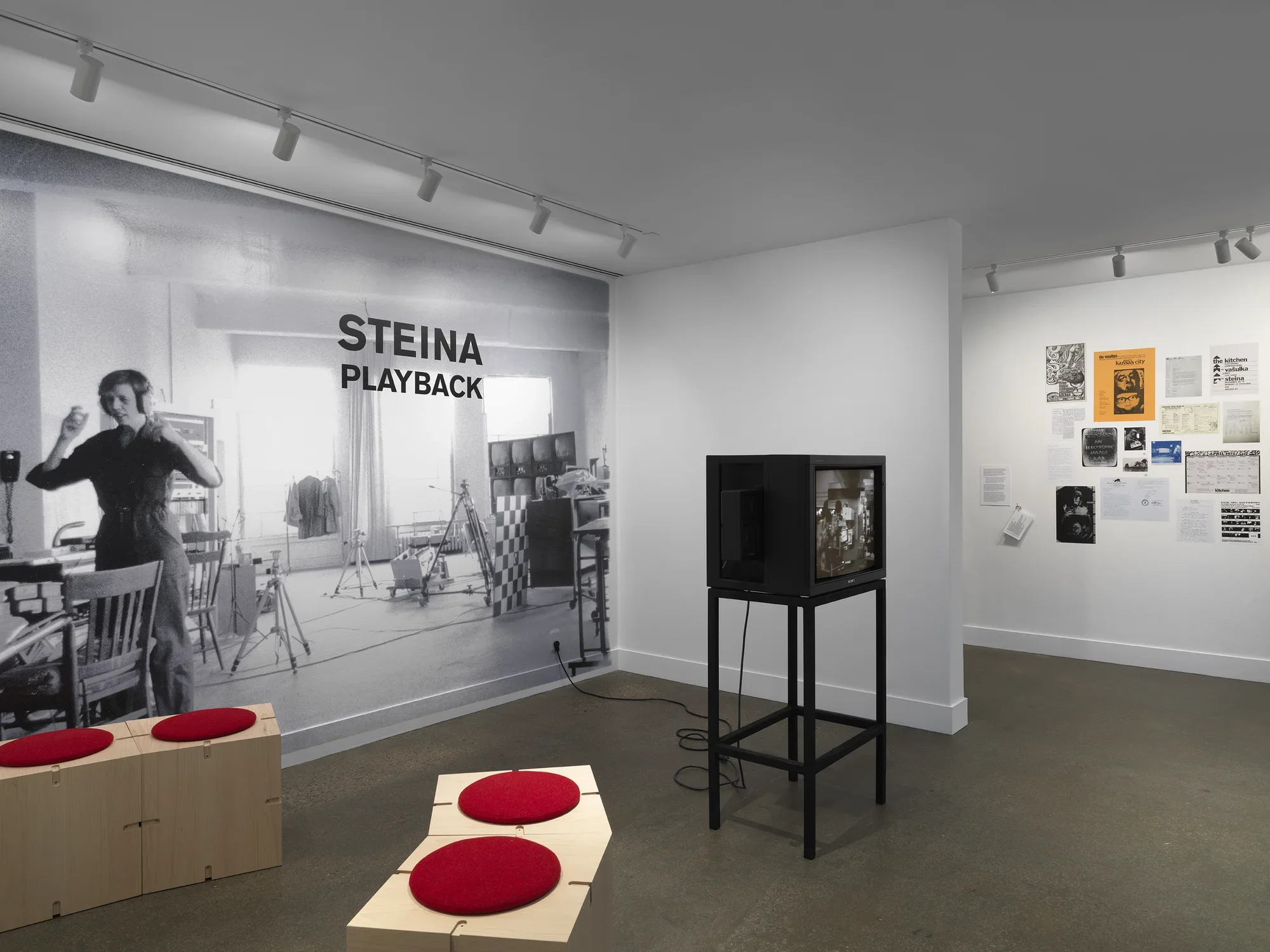 A black-and-white photograph printed mural-size on a wall shows a woman‬ ‭ wearing headphones. She is located in a studio full of technical equipment: tripods,‬ ‭video monitors, wiring. The photograph is overlaid with text indicating the exhibition title:‬ ‭ “Steina: Playback.” The gallery space also includes a boxy video monitor and wooden‬ ‭ seating. In the background on the right, posters and other documents are pasted on the‬ ‭wall.‬ ‭
