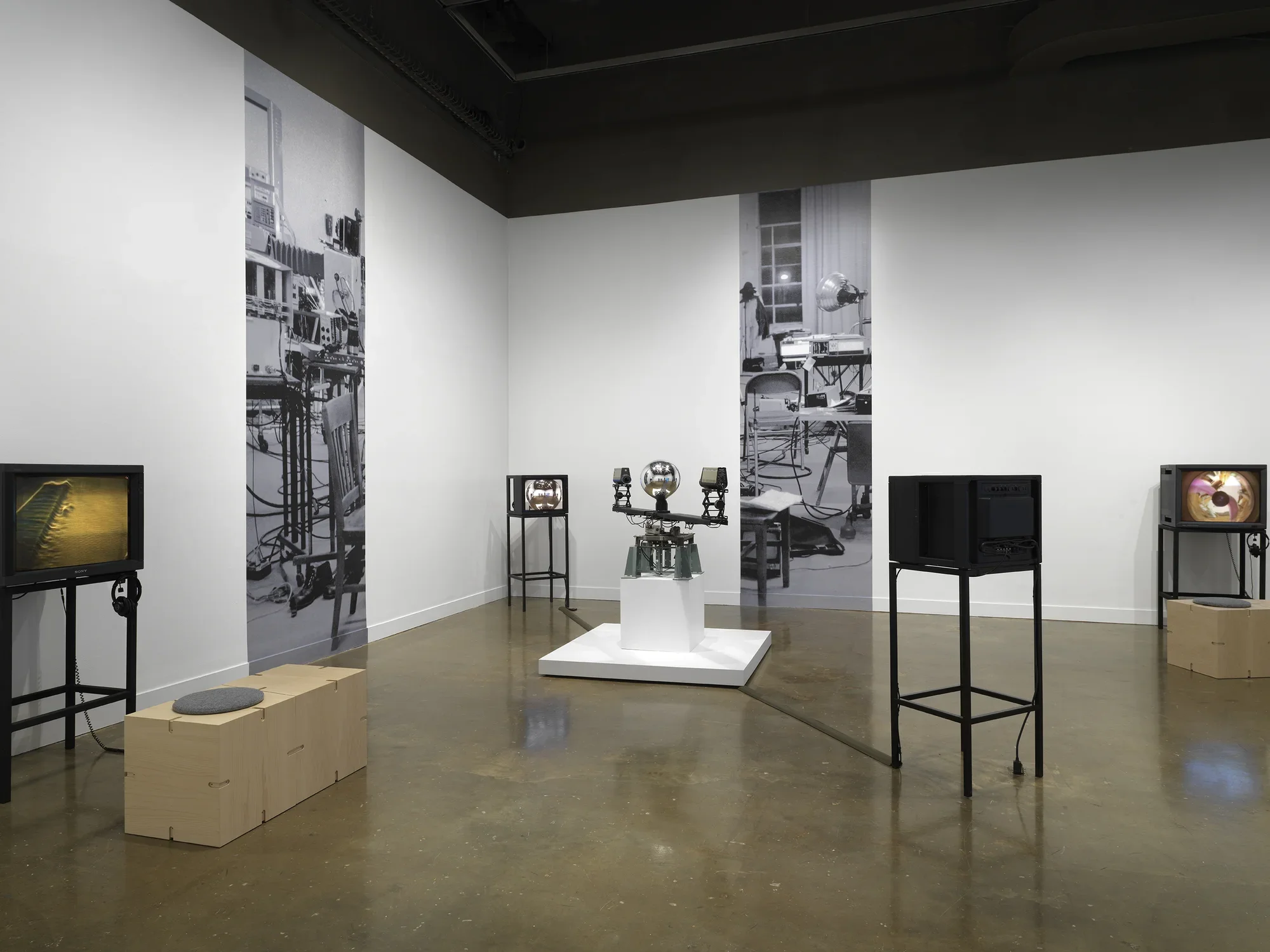 In the center, a sculptural apparatus comprises two cameras on the ends of a‬ ‭ single rotating arm, each pointed inwards at a central mirrored sphere. Feeds from the‬ ‭ cameras are routed to two monitors in the space. On the walls nearly, strips of a‬ ‭ photograph of a studio are pasted on the wall at large scale. On either edge of the‬ ‭ image, video monitors display other works; the one on the right shows a body distorted‬ ‭around a circle, recalling the feed of the orb.‬ ‭