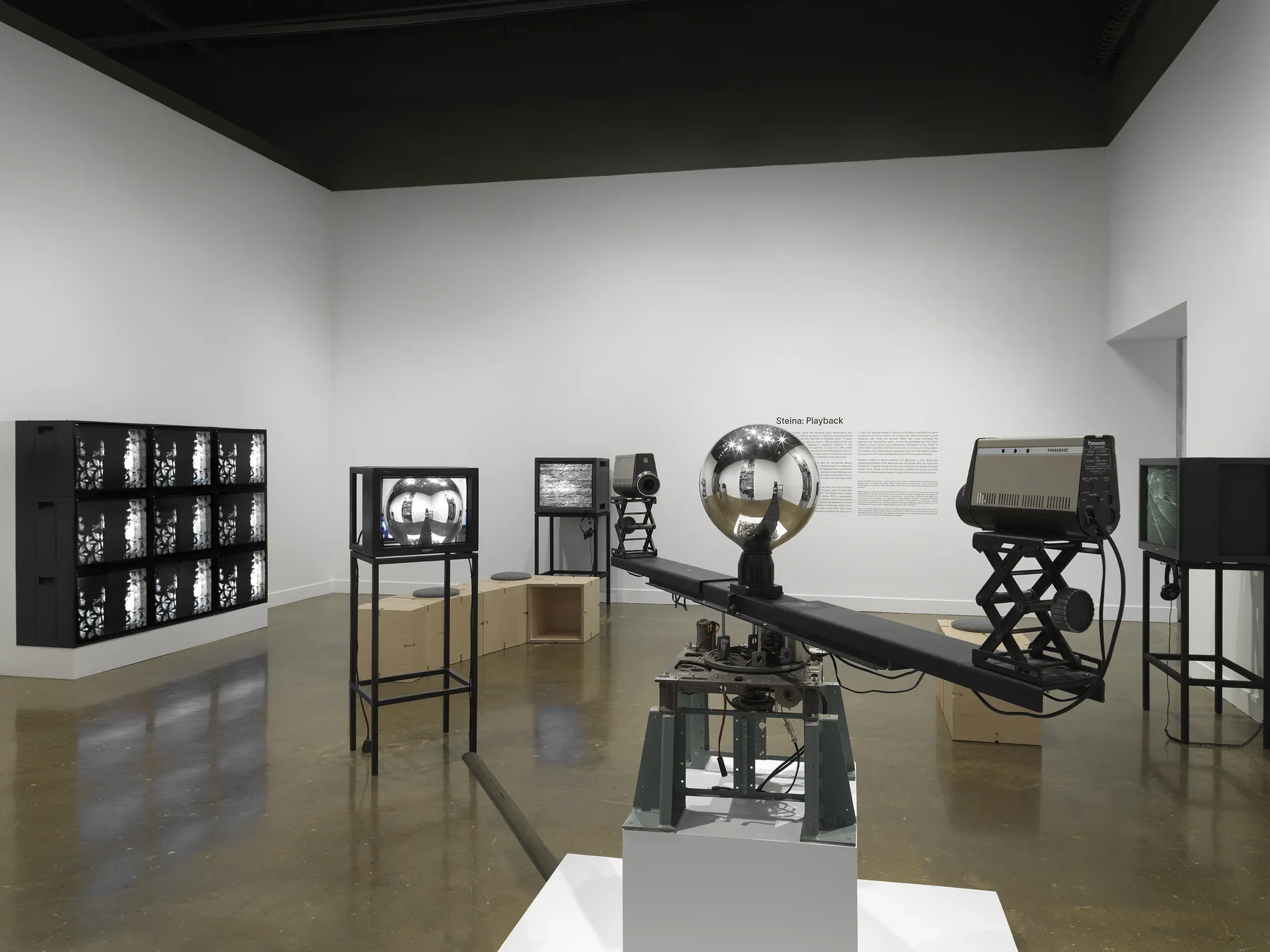 In the foreground, a sculptural apparatus comprises two cameras on the ends‬ ‭ of a single rotating arm, each pointed inwards at a central mirrored sphere. A feed from‬ ‭ the cameras are routed to a nearby monitor. In the background, other video monitors‬ ‭ are visible, including a grid of nine showing the same abstract black-and-white pattern,‬ ‭along with exhibition wall text.‬ ‭