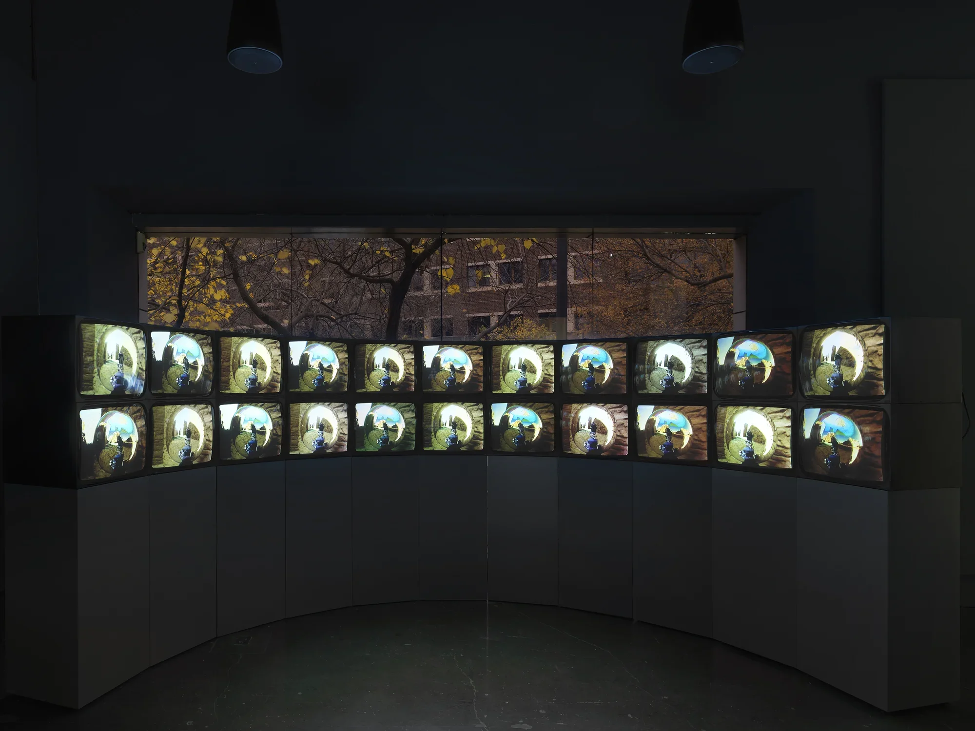 A double-stacked arc of boxy video monitors sits on a pedestal in a darkened‬ ‭ gallery; there are 22 in all. The monitors show similar but not identical images: in each,‬ ‭ a central mirrored orb reflects and distorts the surrounding environment, which includes‬ ‭ a stone wall and blue sky. Behind the video installation is a large window, through which‬ ‭ trees and another building can be seen.‬