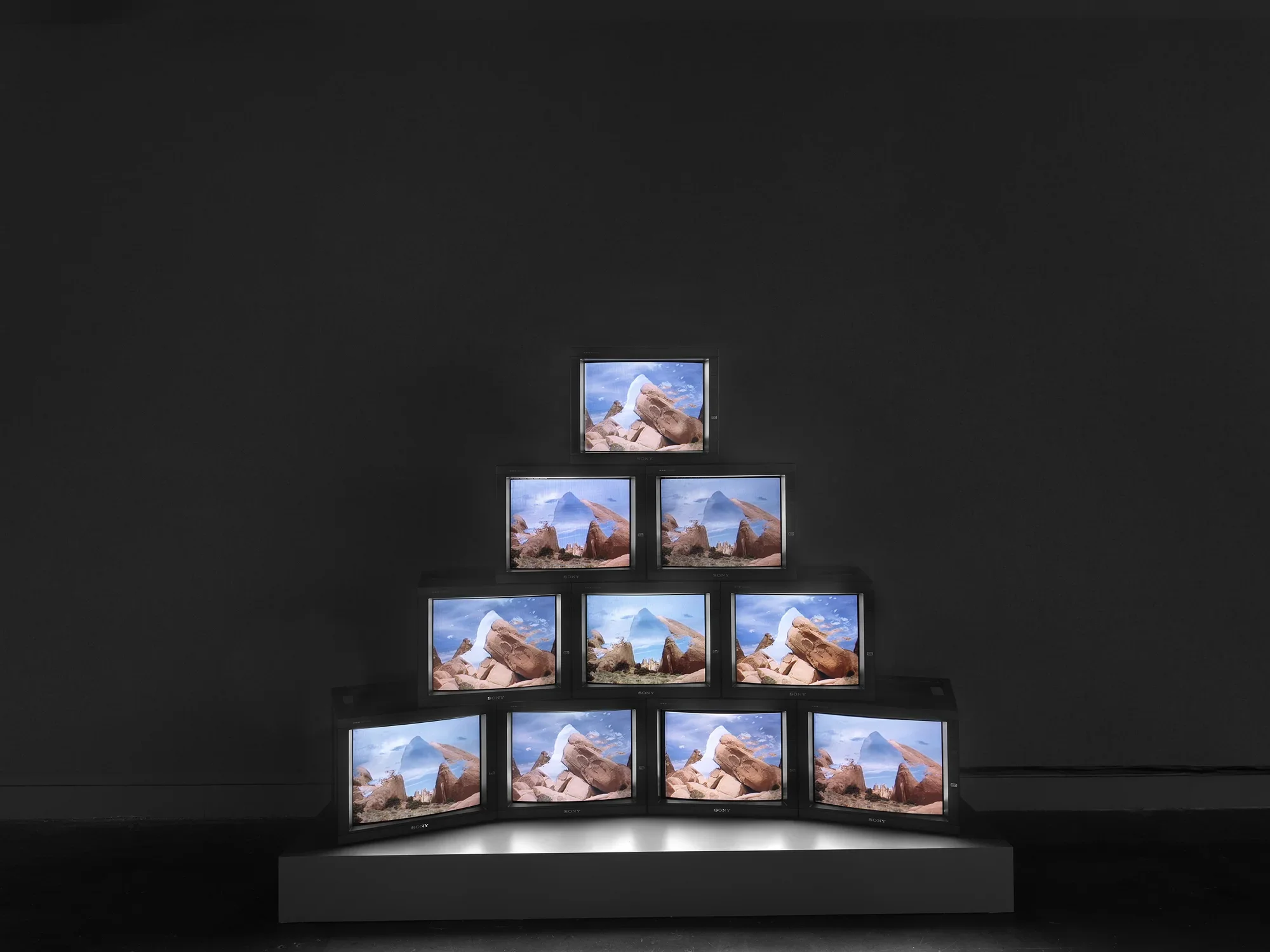 A pyramid of boxy monitors sits on a low pedestal in a dark space. The‬ ‭monitors display images of rocks against blue sky. Closer inspection reveals the images‬ ‭ have been electronically manipulated, overlaying multiple landscapes or sections of sky.‬