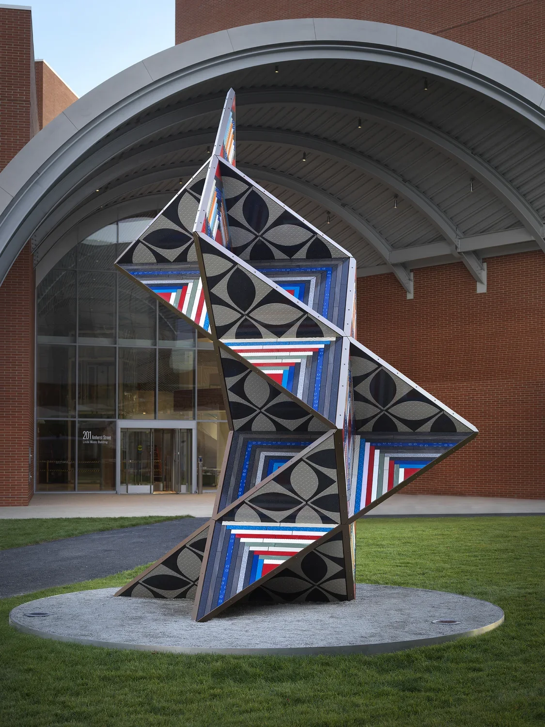 Large brightly colored geometric sculpture in the middle of a grass lawn in front of a brick building with a concrete dome awning.