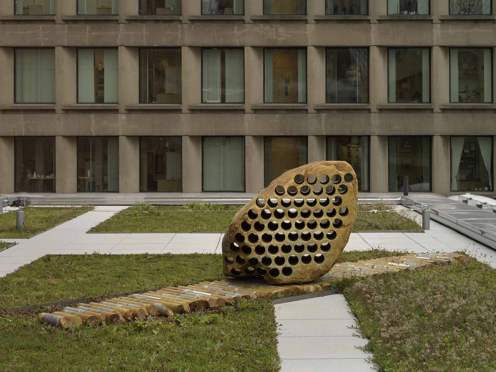 A large rock sculpture rests on a long strip of stone cylinders in a‬ ‭greenspace. The rock has a series of holes removed from its core, with the‬‭ cylinders that were removed making up the strip it rests on. There is a pathway behind the sculpture and building tops surrounding the open space.