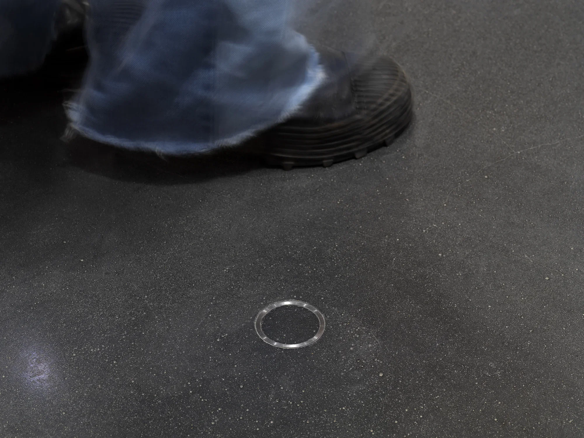 A small metal ring is cemented into the floor. Someone wearing a black sneaker and blue jeans is walking behind the metal ring.