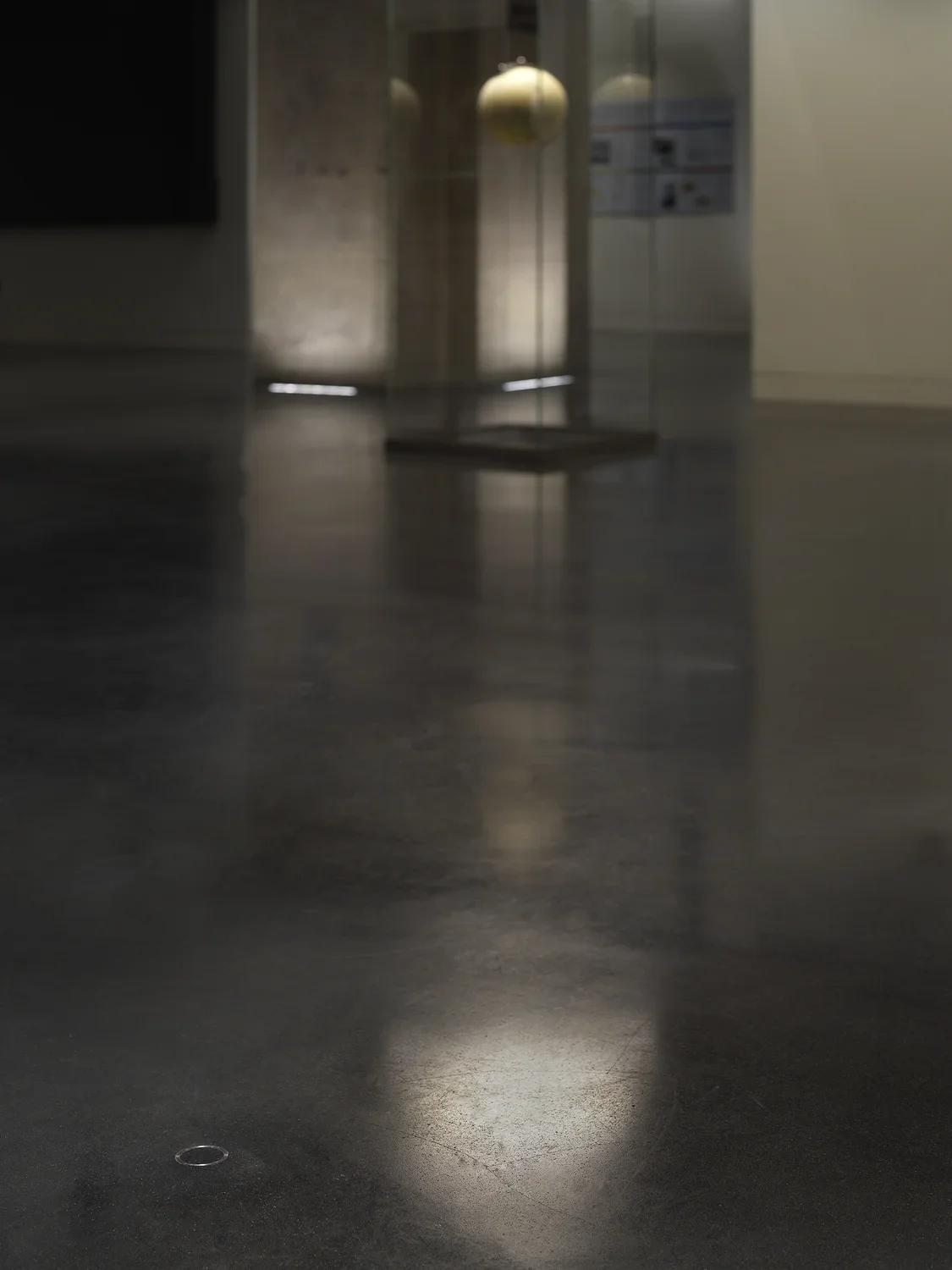 In the bottom left corner of the image, a small metal ring is cemented into the floor. In the background, there is a rectangular glass case with an illuminated hanging globe.