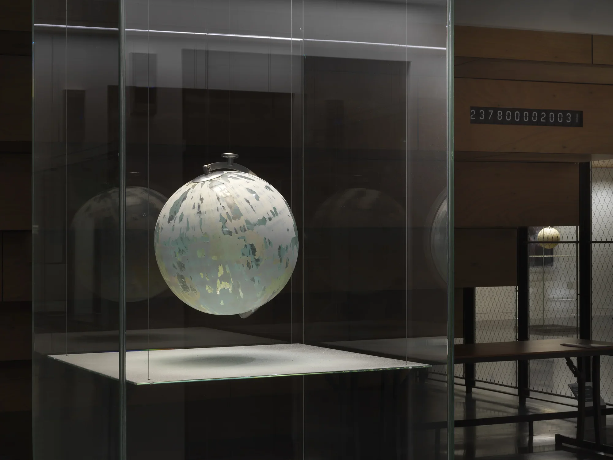 One painted globe suspended in a glass rectangular column and hangs over a flat surface that cuts the cylinder horizontally. Splatters of paint can be seen on top of the panel underneath the globe.