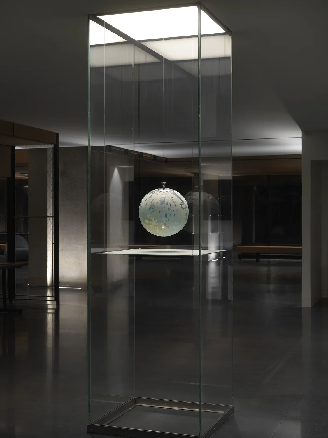 One painted globe suspended in a glass rectangular column and hangs over a flat surface that cuts the cylinder horizontally. Splatters of paint can be seen on top of the panel underneath the globe.