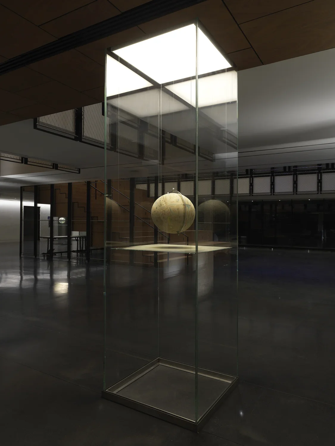 One painted globe suspended in a glass rectangular column and hangs over a flat surface that cuts the cylinder horizontally. Splatters of paint can be seen on top of the panel underneath the globe.