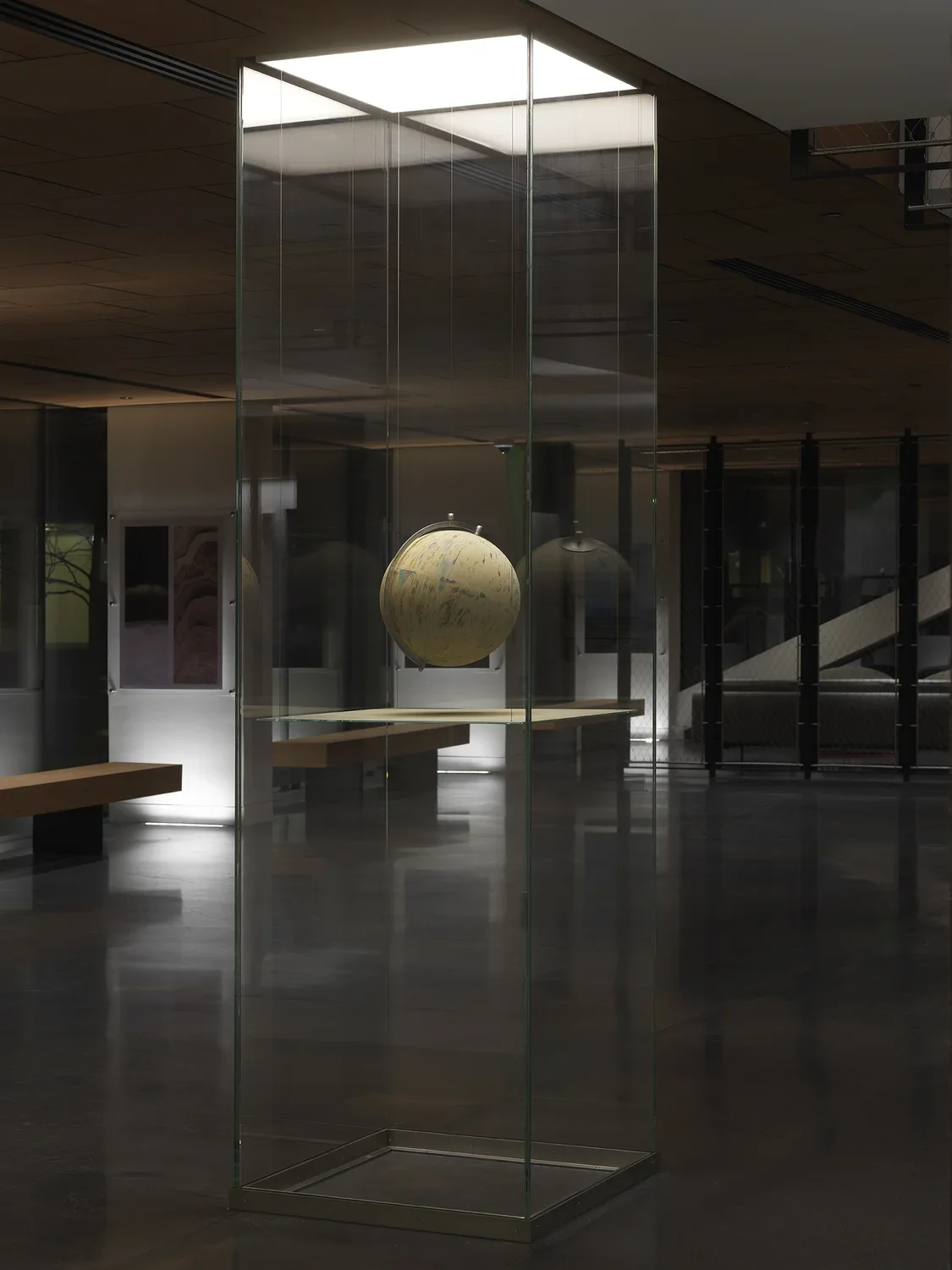 One painted globe suspended in a glass rectangular column and hangs over a flat surface that cuts the cylinder horizontally. Splatters of paint can be seen on top of the panel underneath the globe.