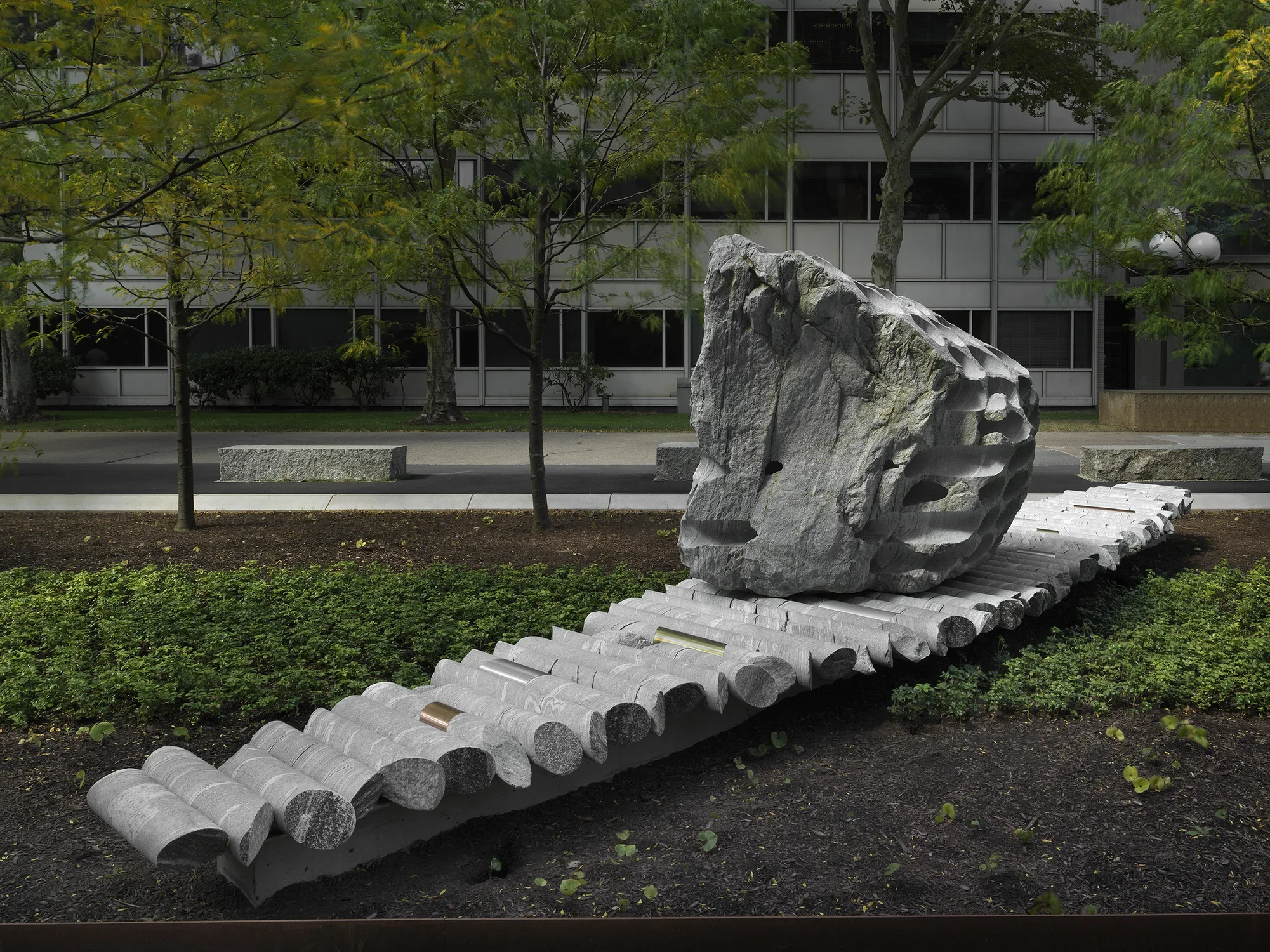 A large rock sculpture rests on a long strip of stone cylinders in a‬ ‭ greenspace. The rock has a series of holes removed from its core, with the‬ ‭ cylinders that were removed making up the strip it rests on. Trees, a pathway,‬ ‭ and a gray building reside in the background.‬
