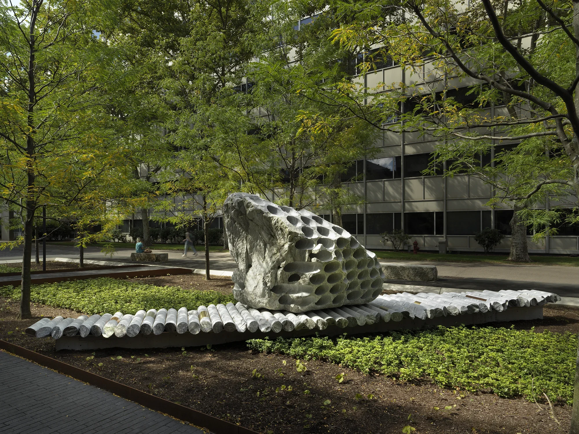 A large rock sculpture rests on a long strip of stone cylinders in a‬ ‭ greenspace. The rock has a series of holes removed from its core, with the‬ ‭ cylinders that were removed making up the strip it rests on. Trees, a pathway,‬ ‭ and a gray building reside in the background.‬
