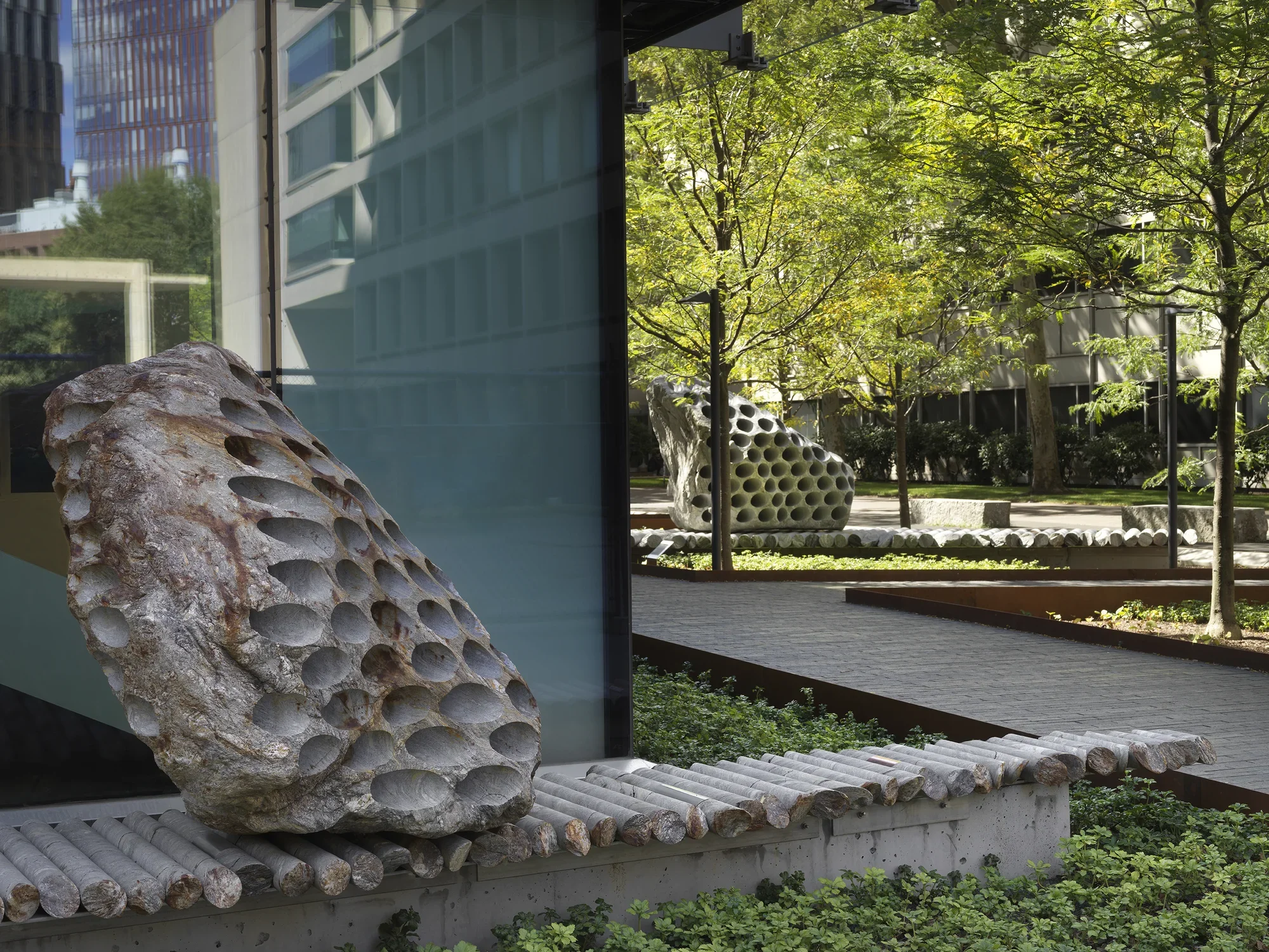 Two large rock sculptures rest on long strips of stone cylinders in a green space next to the corner of a building. each rock has a series of holes removed from its core, with the cylinders that were removed making up the strip it rests on. One sculpture is located in the lower left corner of the image, with the other in the background surrounded by trees.