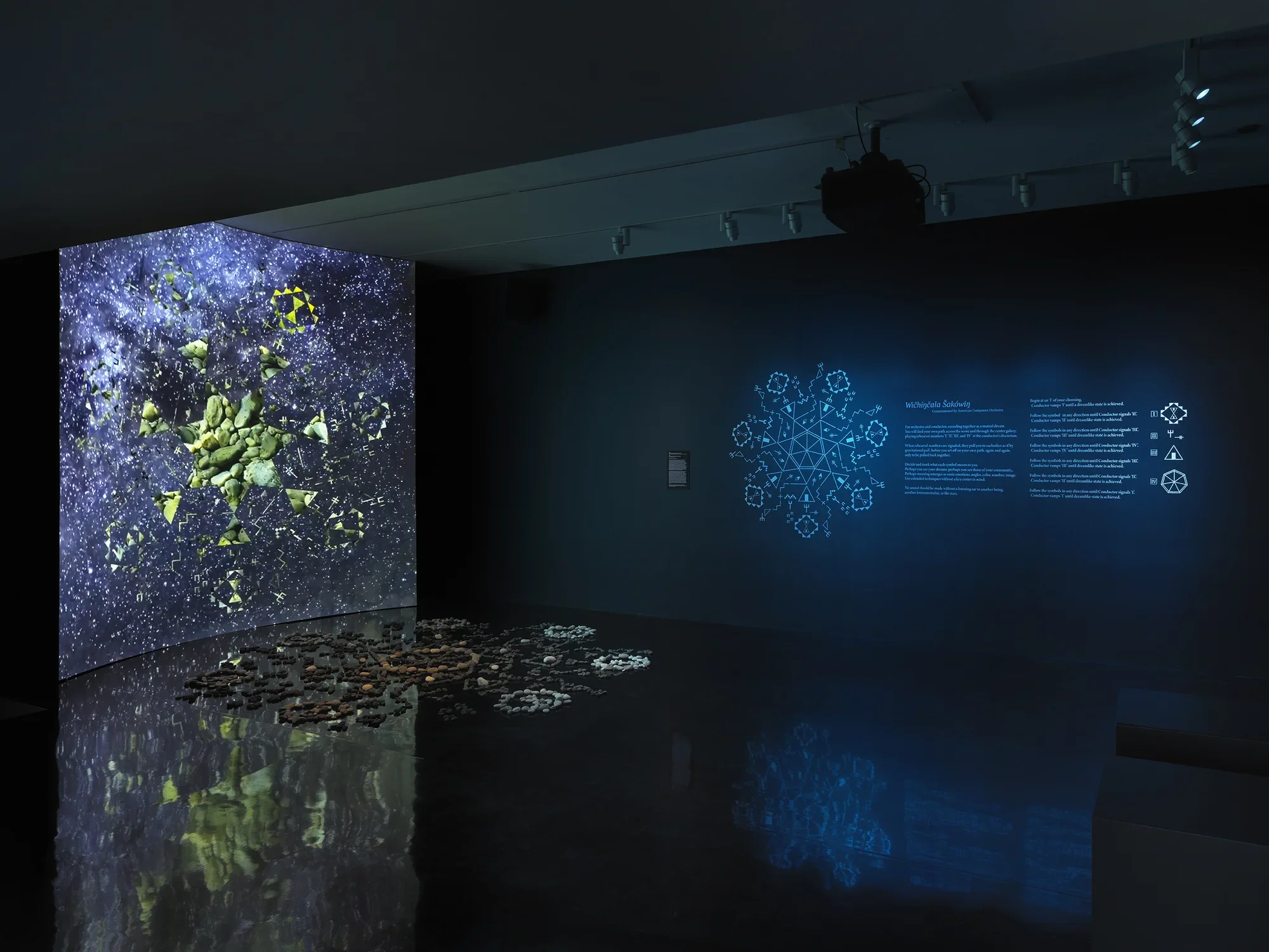 A dark gallery space bathed in blue light. On the left, a large screen displays‬ ‭ a six-sided star at its center, within which video footage of green stones is projected.‬ ‭ Surrounding the star, six inward-facing triangles form a pattern. The background‬ ‭ blends swirling geometric shapes with footage of stars and the night sky. In front of‬ ‭ the screen, a formation of stones rests on the reflective black floor. On the right wall,‬ ‭glowing blue geometric forms and wall text are visible.‬ 