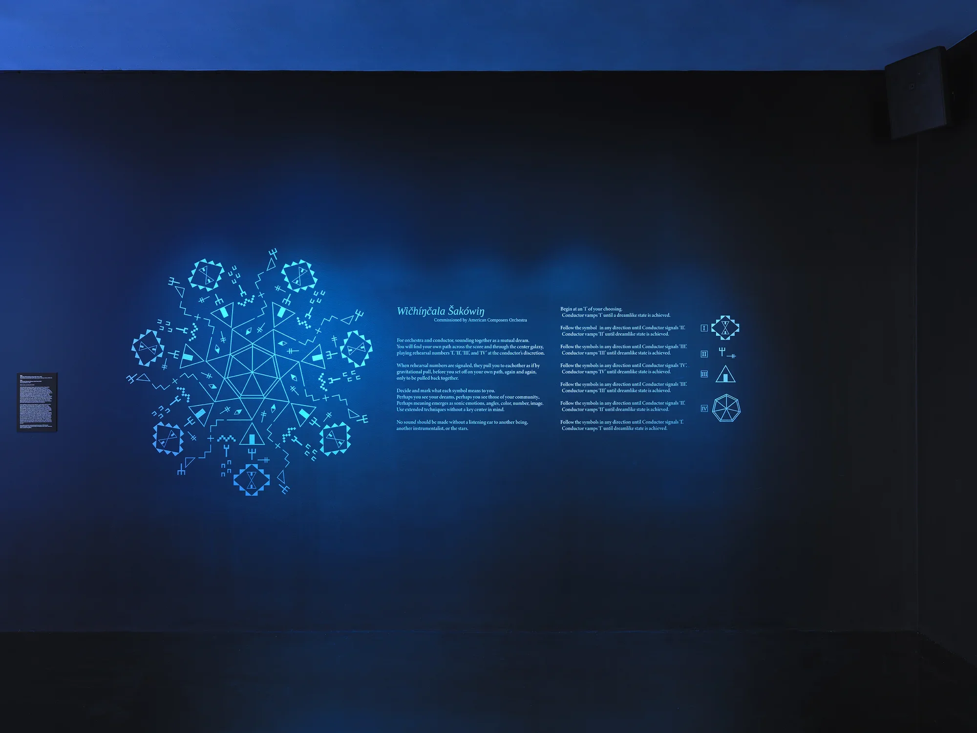 On a dark blue gallery wall, a glowing circle of geometric forms made up of‬ ‭ Lakhota characters is displayed. To its right, instructions for a score commissioned‬ ‭ by the American Composers Orchestra are present. Further to the right, four‬ ‭ Lakhota characters are shown, representing the elements that compose the‬ ‭geometric form on the left.‬ 