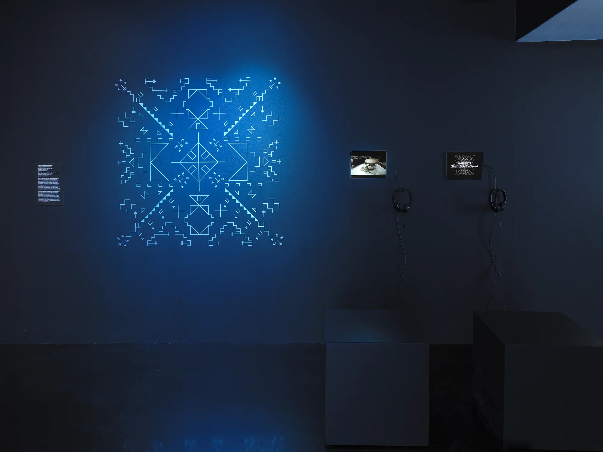 In a dark gallery space, a blue-glowing geometric form is shown on the wall.‬ ‭To its right, two small iPads showcase video footage—one featuring a drum and the‬ ‭other displaying a musical score.‬ 