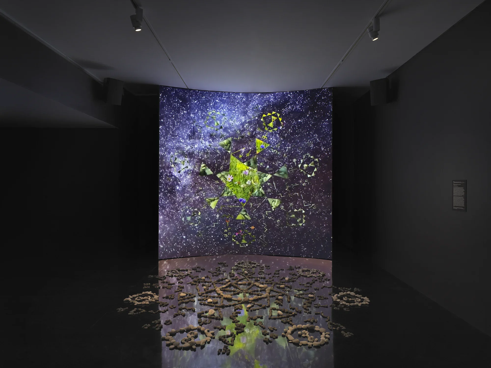 A dark gallery space features a large screen displaying a six-sided star at its‬ ‭ center. Within the star, video footage of green stones is projected. Surrounding the‬ ‭ star, six inward-facing triangles form a pattern, while geometric shapes swirl in the‬ ‭ background alongside footage of stars and the night sky. In front of the screen, a‬ ‭formation of stones rests on the reflective black floor.‬ ‭