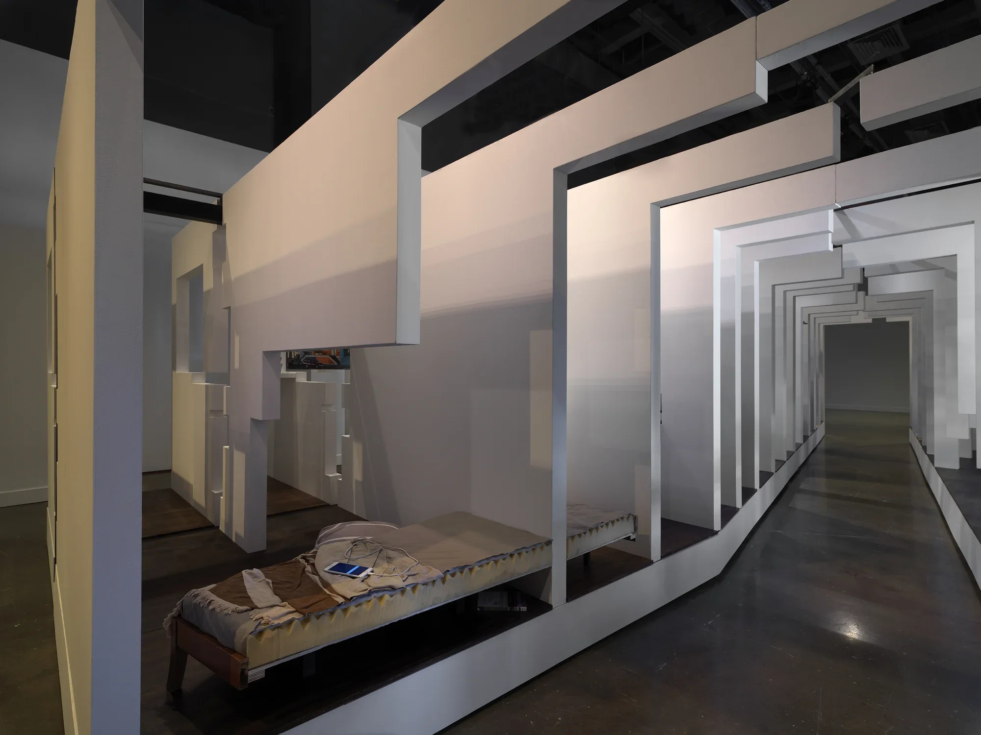 A walkway surrounded on both sides by a series of white walls that segments what appears to be a living space. A charging smartphone is visible on top of a bed in the bottom lefthand corner.