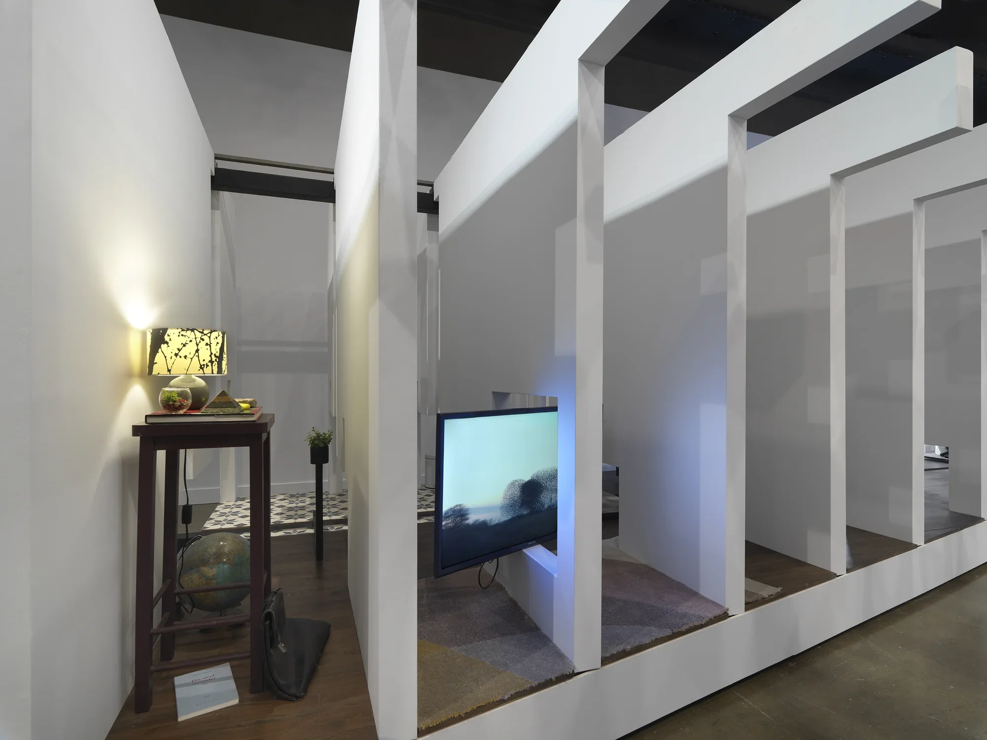 A series of fragmented white walls divides a living space with wooden floors, furniture, and house plants throughout. Each section of wall is connected by black beams. There is a wooden nightstand with a lamp on top and a globe on the ground beside at the left side of the frame. A flatscreen TV is divided by a white wall on the right side of the frame.