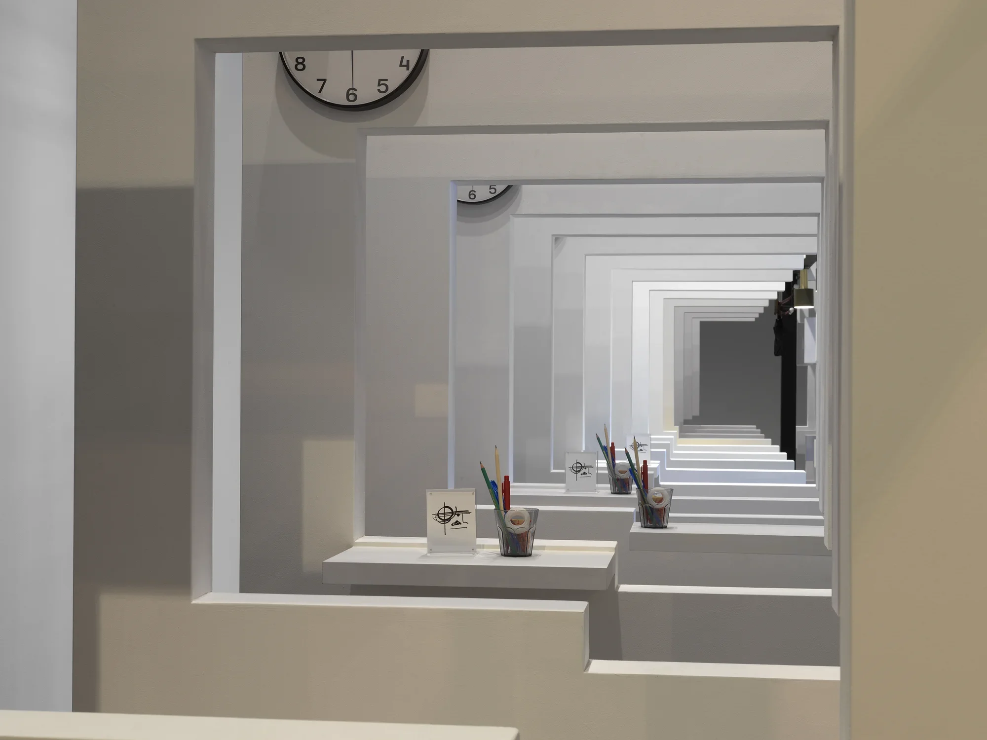 A series of white walls with a window cutout that has a view of the next white wall with a window cutout. There is a cup with office supplies on the window ledge of each wall.