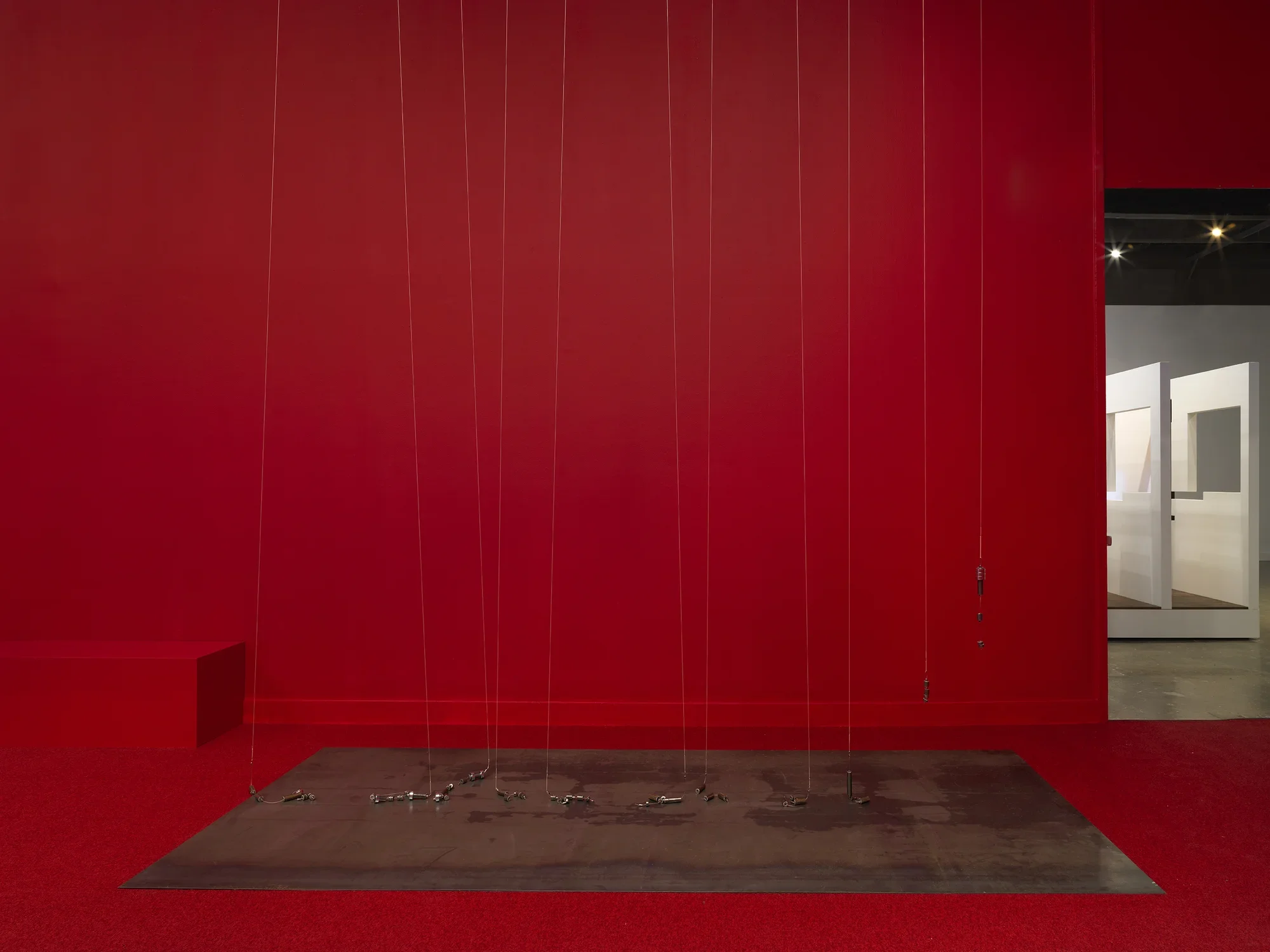 A series of weights suspended on strings above a metal plate in the middle of a completely red painted walled room with red carpeting. On the right side there is a doorway to bright room.
