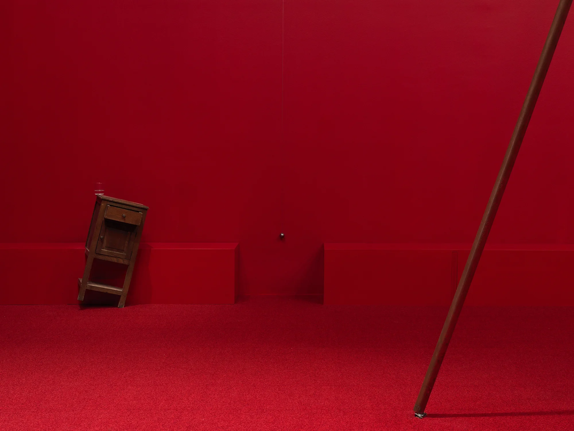 A wooden cabinet standing on a angle tilted to the right in a completely red painted room with red carpeting, next to a metal sphere hanging on a string suspended from the ceiling. Part of a copper poll is angled to the right in the right hand side of the frame.