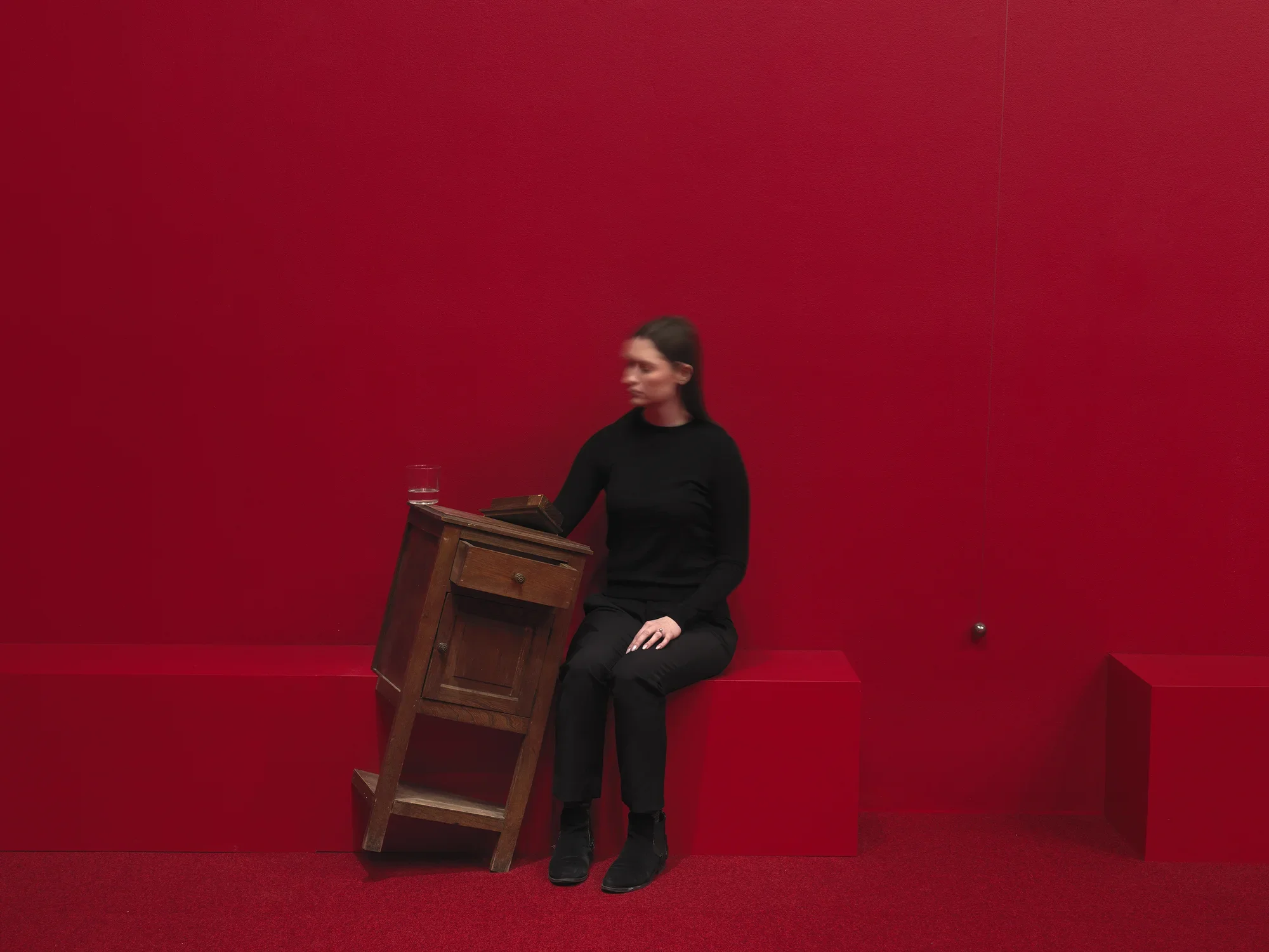 A wooden cabinet tilted on a angle to the right in a completely red painted room with red carpeting. A woman in a black sits to the right with her hand inside of the cabinet. A swinging metal sphere on a string suspended from the ceiling is captured in motion.