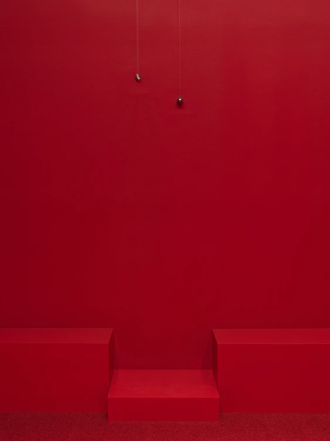 Two swinging metal spheres suspended from the ceiling on strings are captured in motion in a completely red painted walled room with red carpet.
