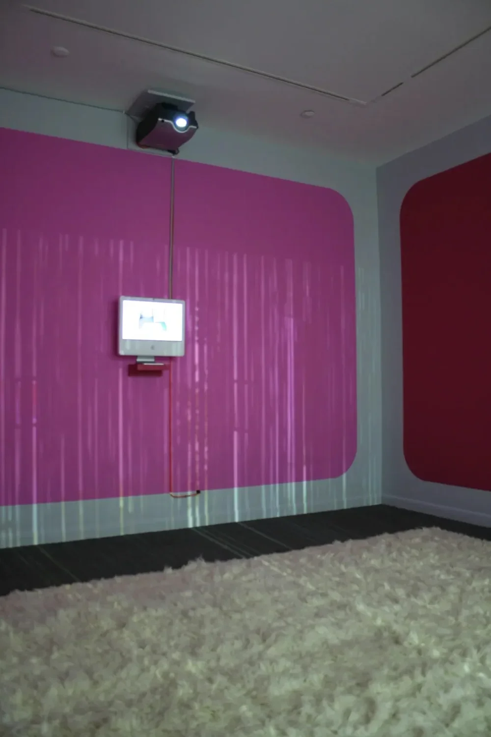 2007 Mac computer monitor affixed to a wall with a pink rectangle with a plush white rug on the ground. 