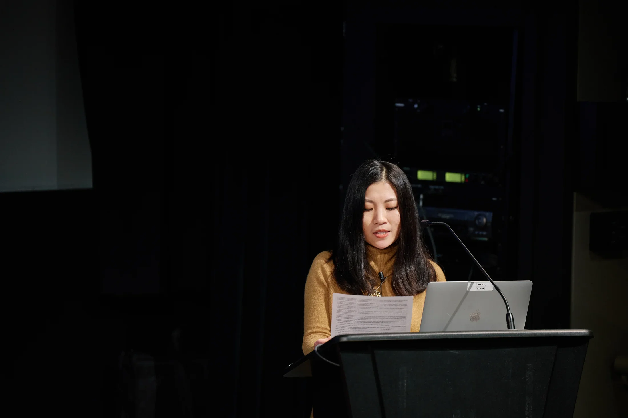 A woman with long dark hair is reading from a stack of paper while behind a podium with a tall microphone and a silver Mac laptop. The background is dark behind her except for two green squares.