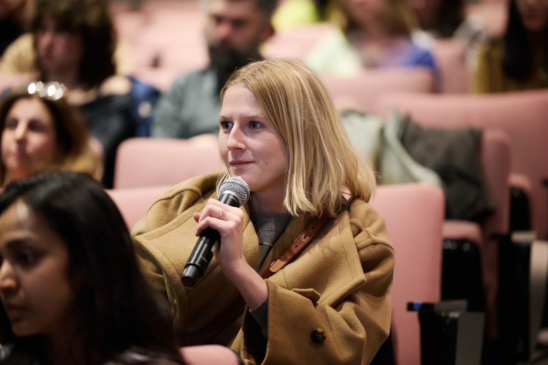 Woman with short blonde hair in a camel colored jacket sitting in a theater audience, holding a microphone near her mouth.