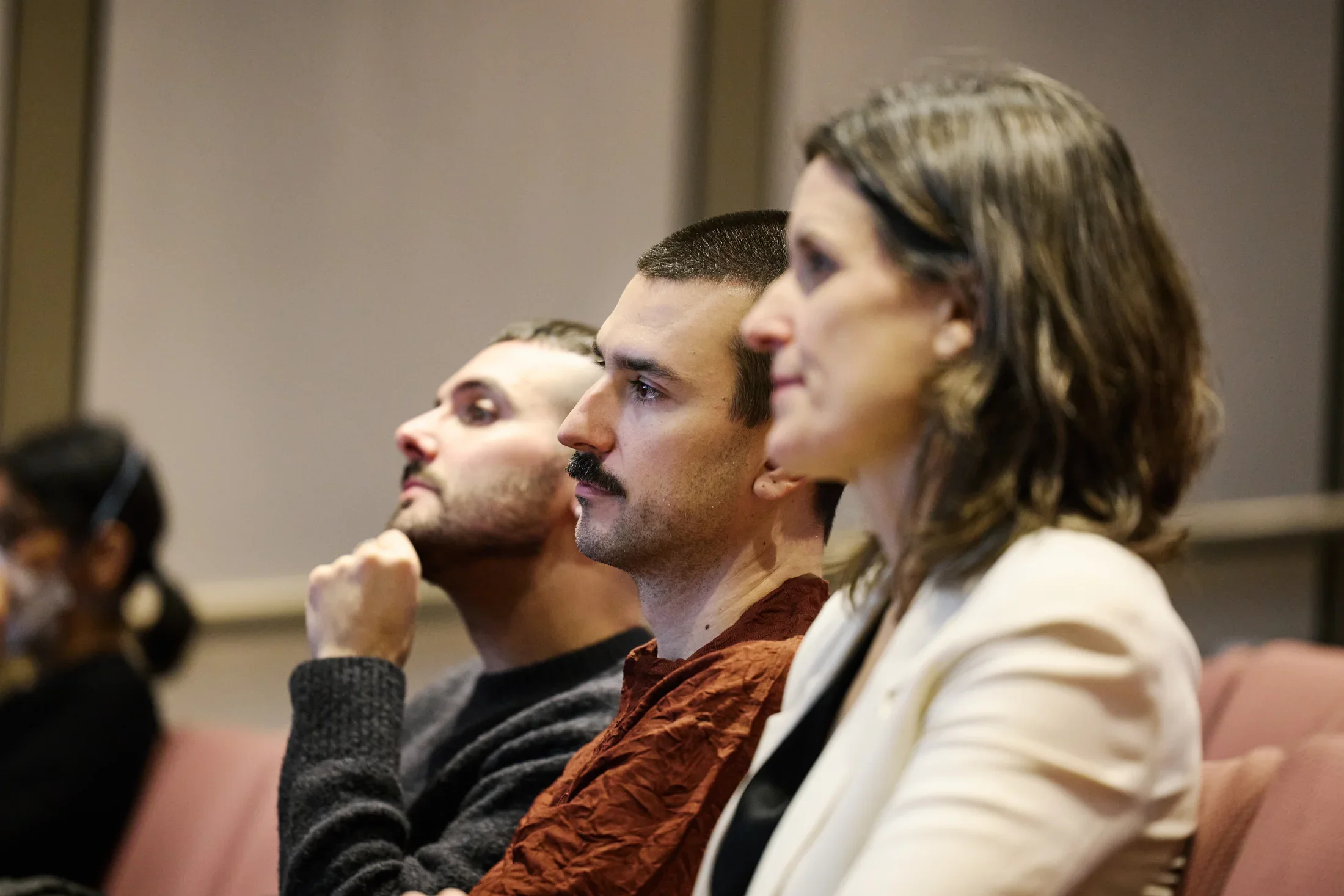 Three people sitting in red theater chairs face the left side of the image, looking at something out of the frame. The person furthest from the camera has their hand on their chin inquisitively.