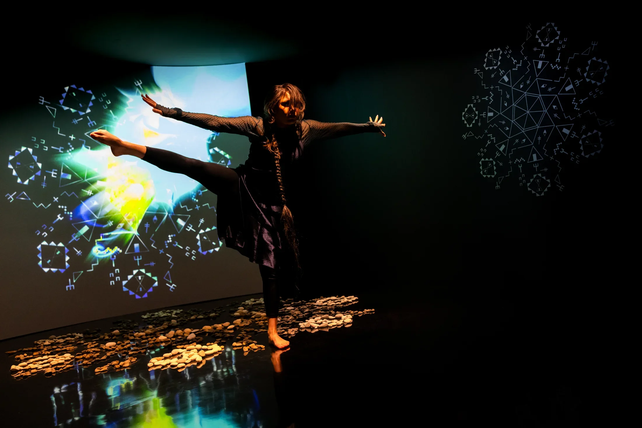 Still of a performance showcasing a woman spotlighted on stage with her arms out to both sides facing the camera and her right leg raised to hip height with foot pointed. A brightly colored geometric pattern in the background is juxtaposed by a black wall.