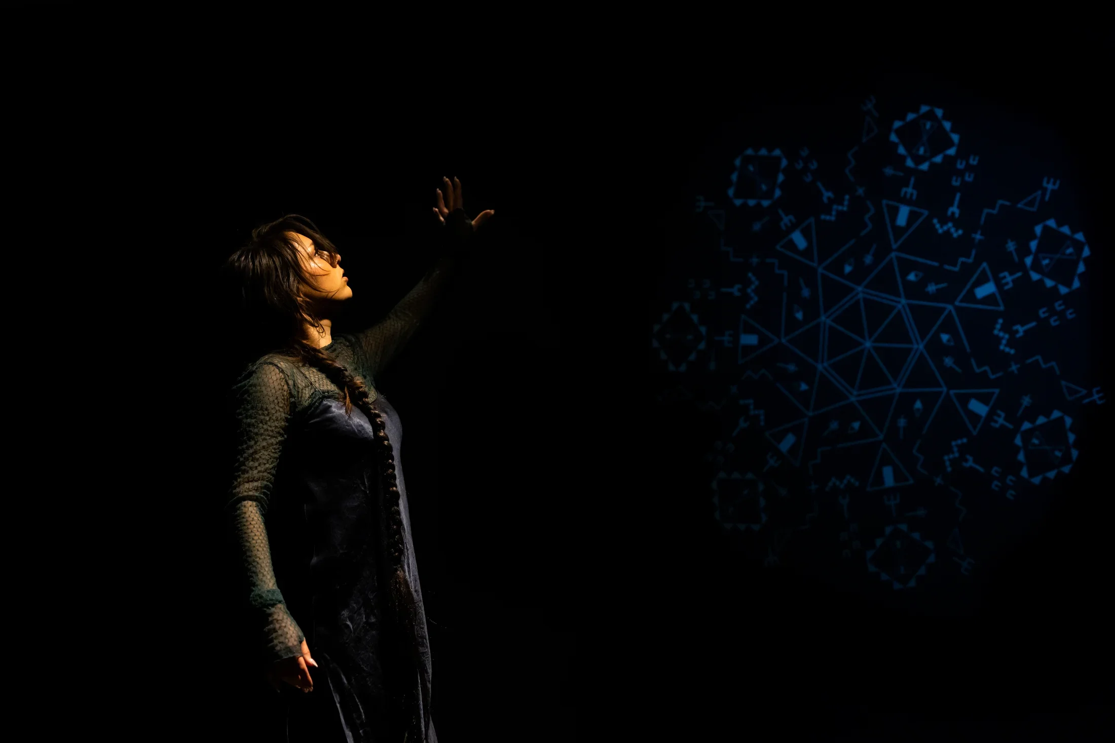 Still of a performance showcasing a woman spotlighted on stage with her left arm raised above her head. A faint colored geometric pattern in the background is juxtaposed by a black wall.