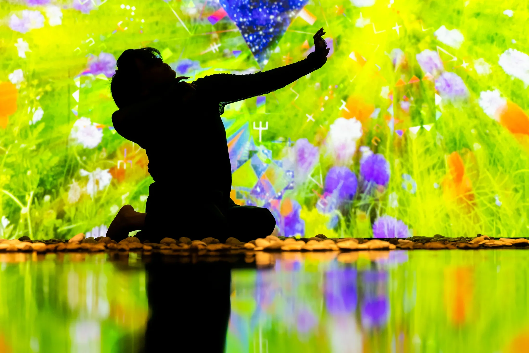Still of a performance showcasing the silhouette of a woman on stage with her arms out to the left side. A brightly colored geometric pattern in the background is reflected on a mirrored floor.