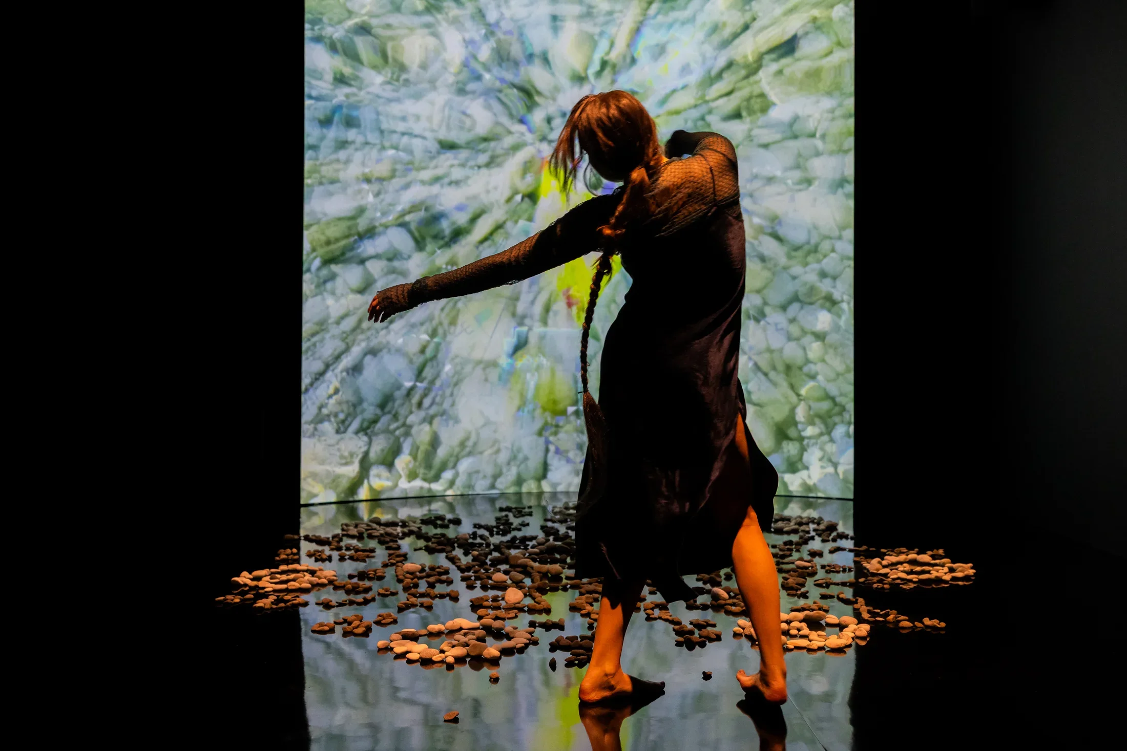 Still of a performance showcasing a woman spotlighted on stage with her arms out to the left side. A brightly colored geometric pattern in the background is juxtaposed by a black wall.